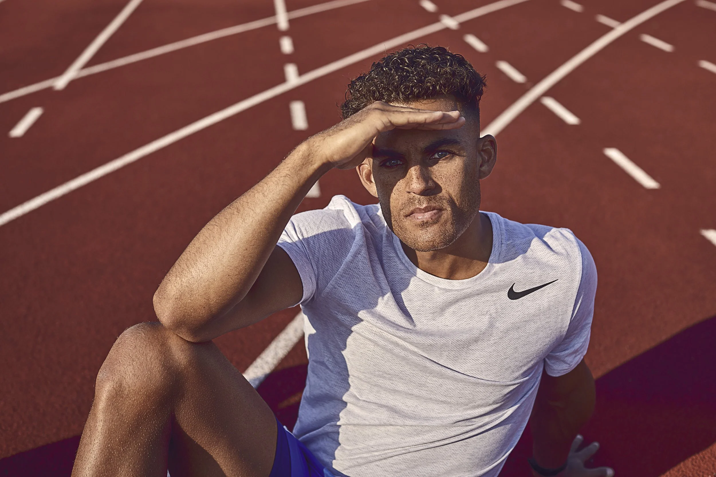 A male athlete sitting on a running track, shielding his eyes from the sun with his hand, wearing a white Nike t-shirt and blue shorts, with white track lines visible in the background.