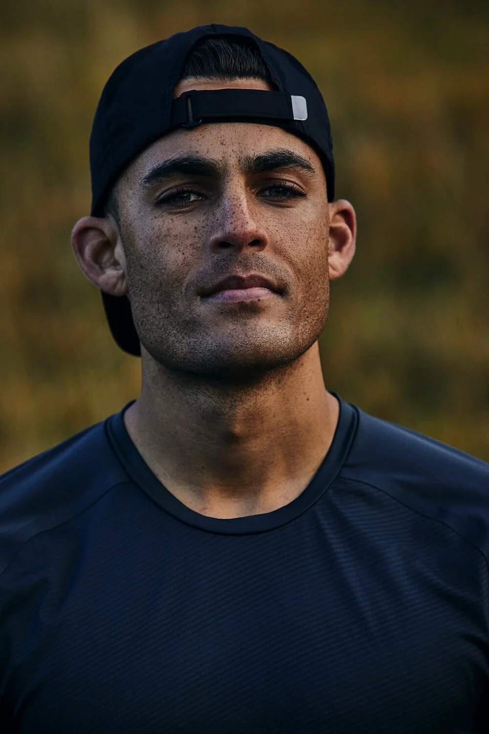 Close-up of a young man wearing a black baseball cap backwards and a black athletic shirt, outdoors during golden hour.