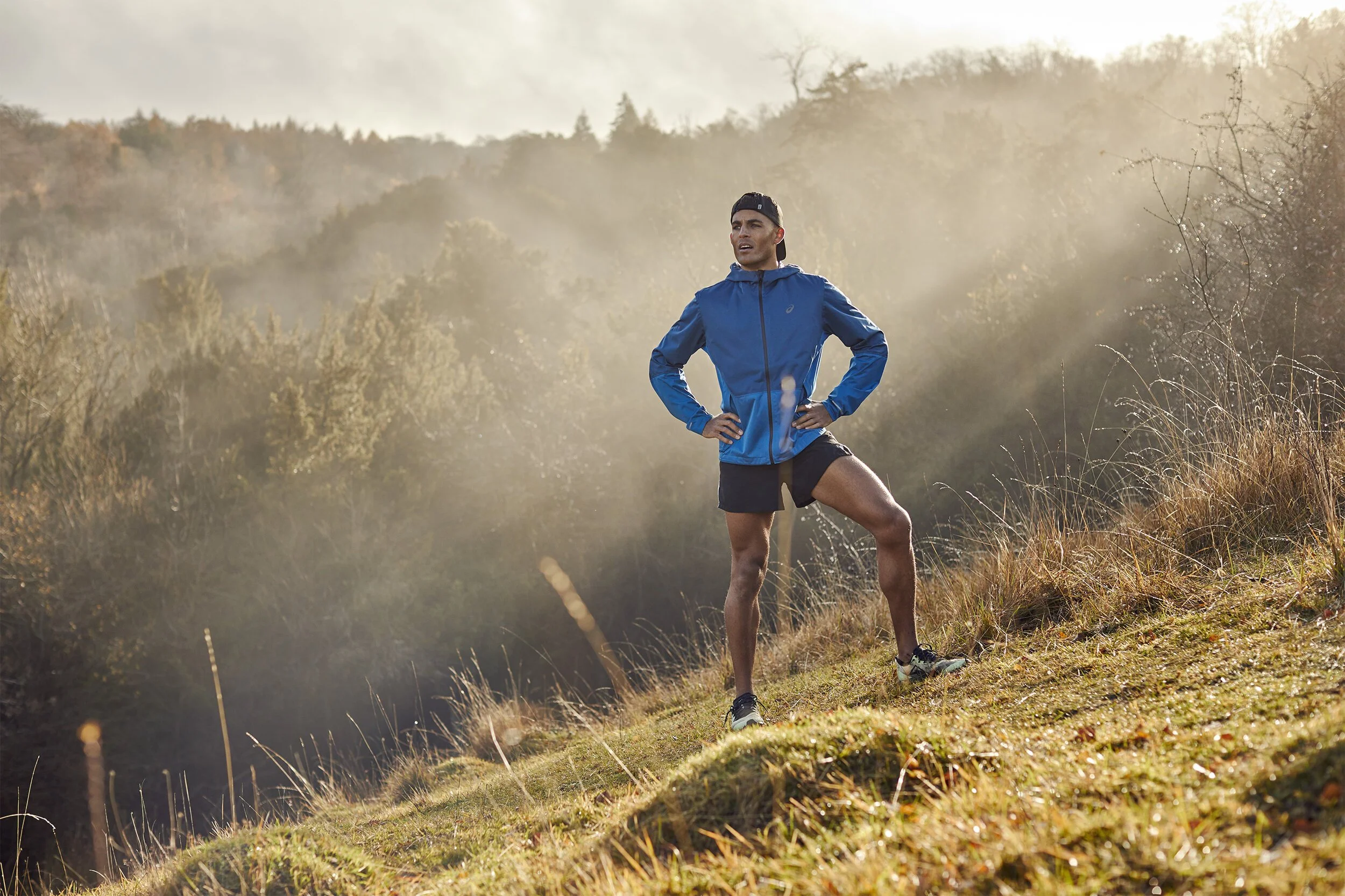 A man in a blue jacket and black shorts standing on a grassy hillside with hands on hips, looking into the distance with a natural landscape background and soft sunlight.