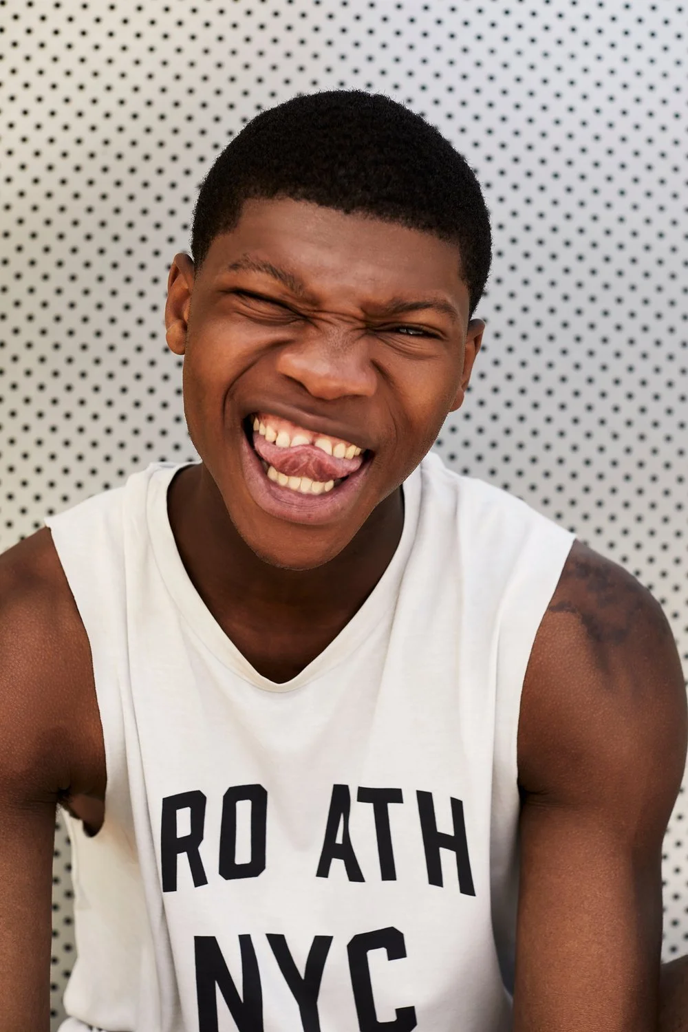 A young man with dark skin and short black hair making a funny face, sticking out his tongue and squinting one eye, in front of a gray dotted wall, wearing a white sleeveless shirt.