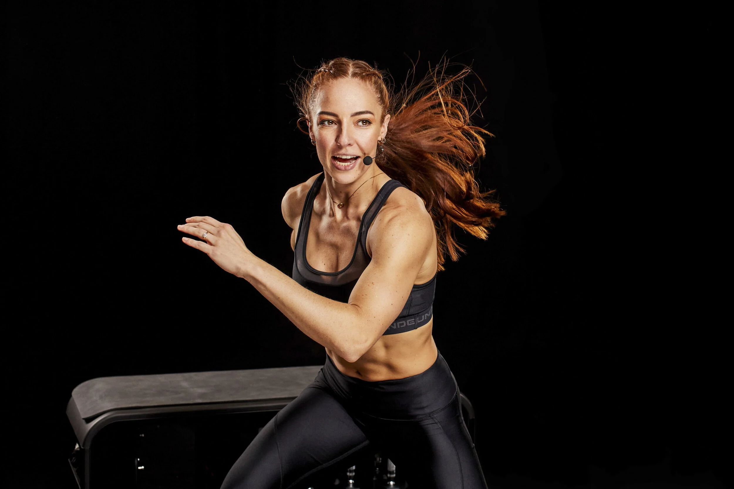 A woman in athletic wear with long, red hair is running on a treadmill against a black background.