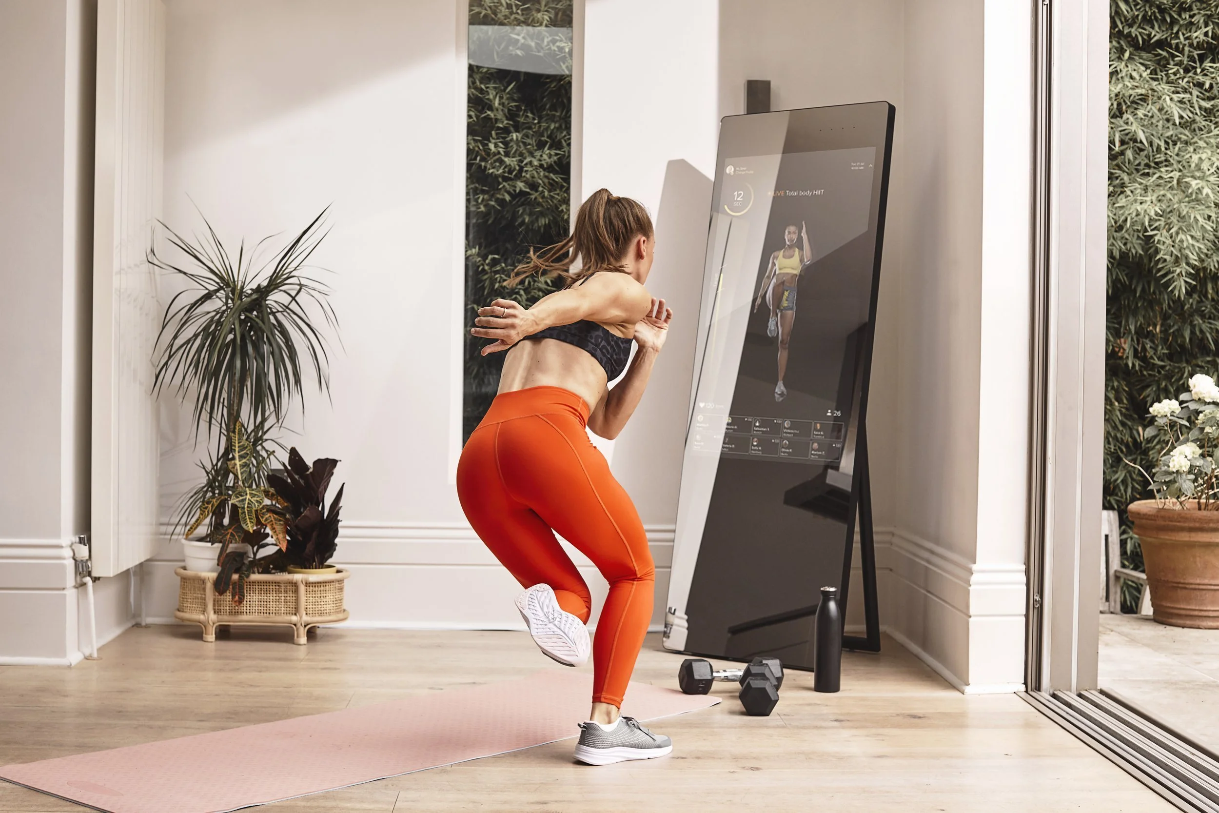 Woman working out on a pink yoga mat with a virtual fitness trainer displayed on a large vertical screen in a bright room with houseplants and sliding glass door.