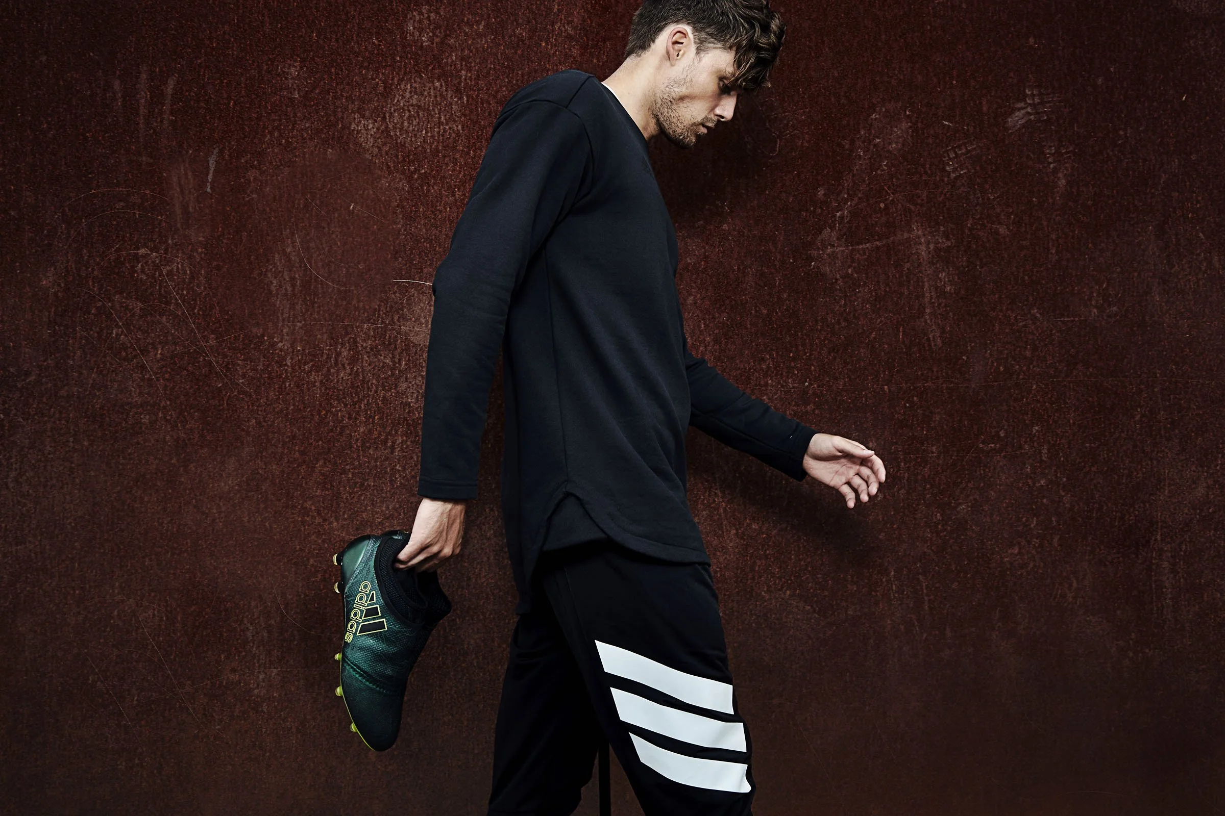 A man stretching, holding a pair of Adidas soccer cleats in his right hand while standing against a textured red wall, wearing black athletic clothing with white stripes.