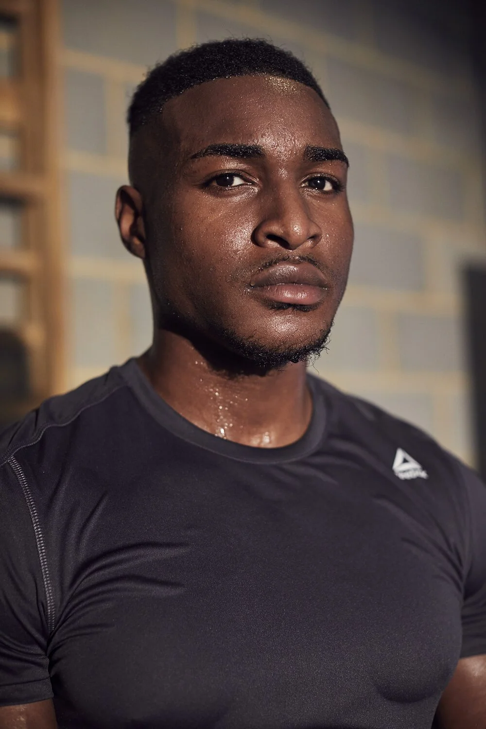 A young man with dark skin and short black hair is seen wearing a black athletic shirt, sweating, in front of a gym wall.