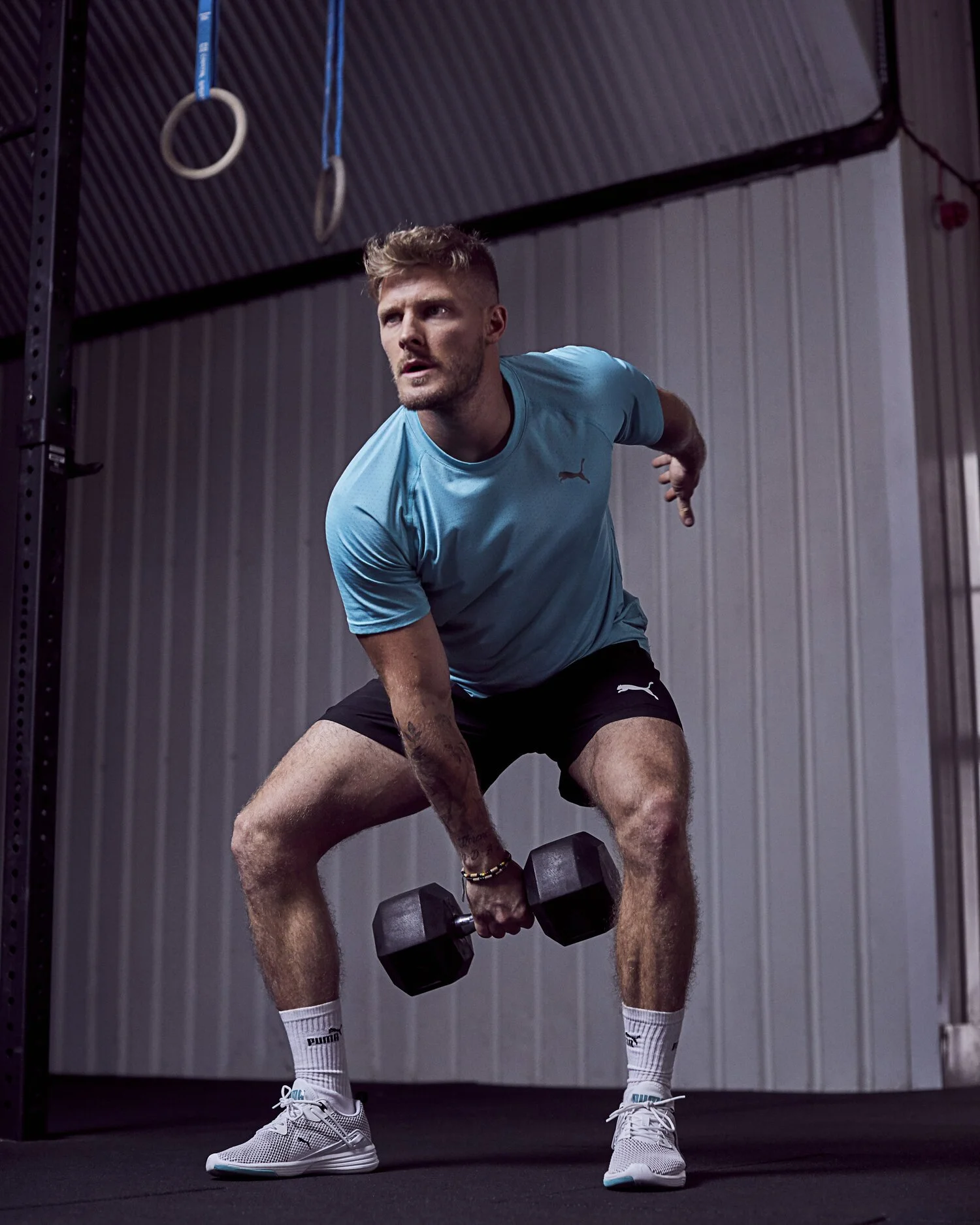 A man in a blue Puma t-shirt and black shorts doing a bent-over dumbbell row exercise in a gym, with gym rings hanging from the ceiling in the background.