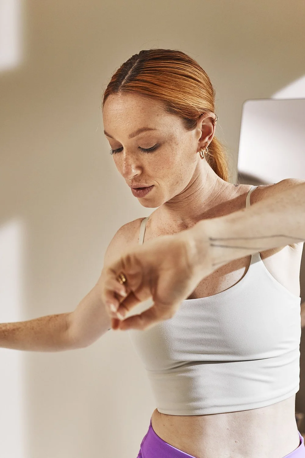 A red-haired woman in a white tank top and purple shorts is applying deodorant to her armpit.