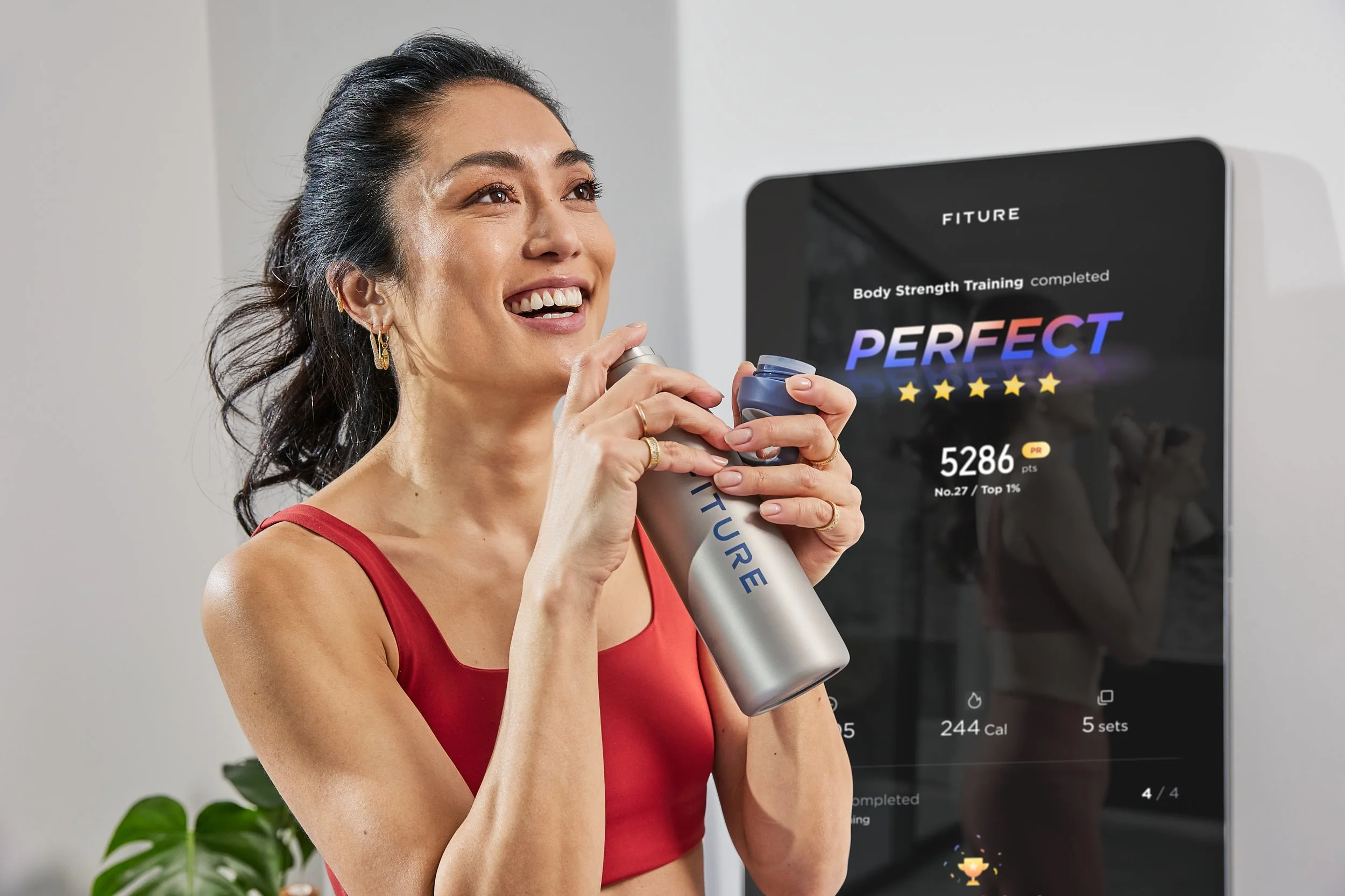 A woman in a red sports bra smiling and holding a water bottle in front of a digital fitness screen showing a workout completion message, calories burned, and points earned.