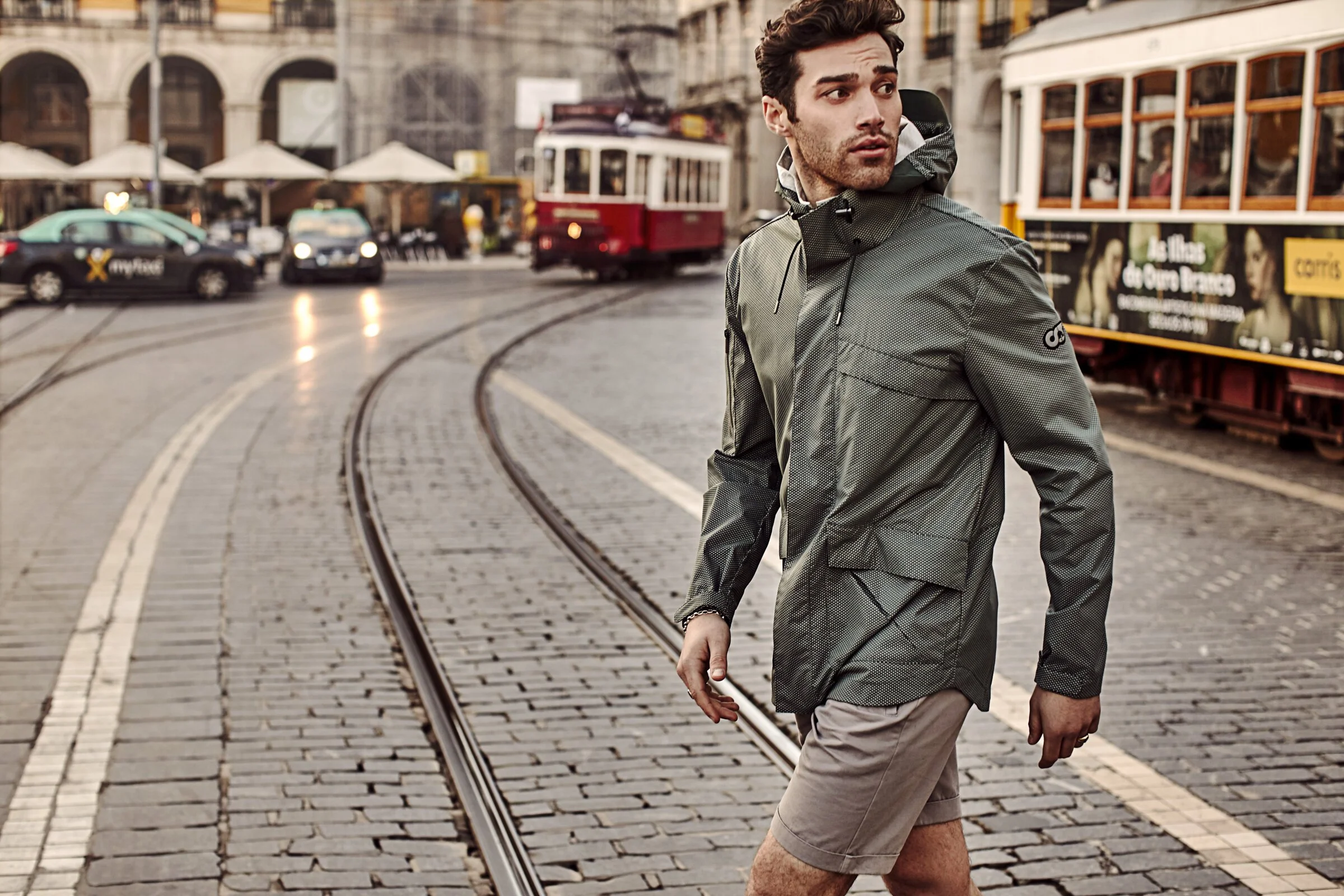 A young man walking on a cobblestone street with tram tracks, wearing a green waterproof jacket and beige shorts, in a city with cars, a tram, and buildings in the background.