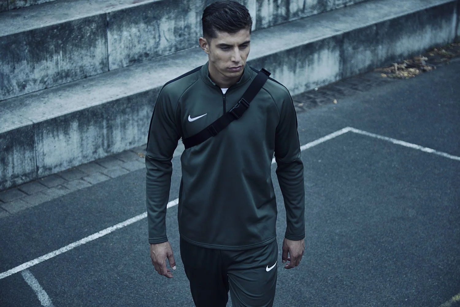 Young man wearing dark gray Nike sportswear and carrying a black strap over his shoulder, walking on a track against a stone wall.