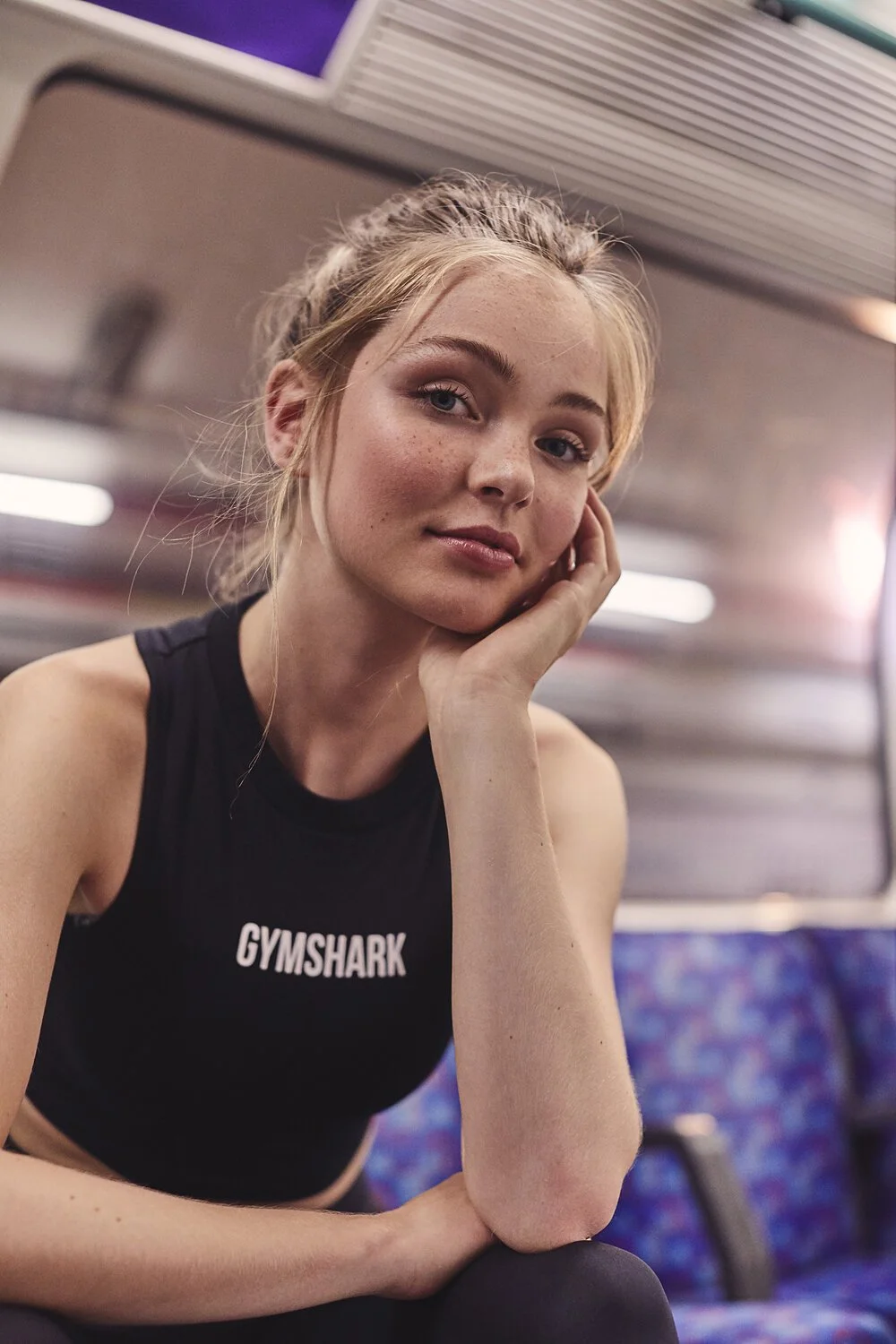 A young woman with blonde hair tied in a bun, wearing a black Gymshark sports top, sitting inside a bus or train with patterned seats and a metallic interior, resting her head on her hand and looking at the camera.