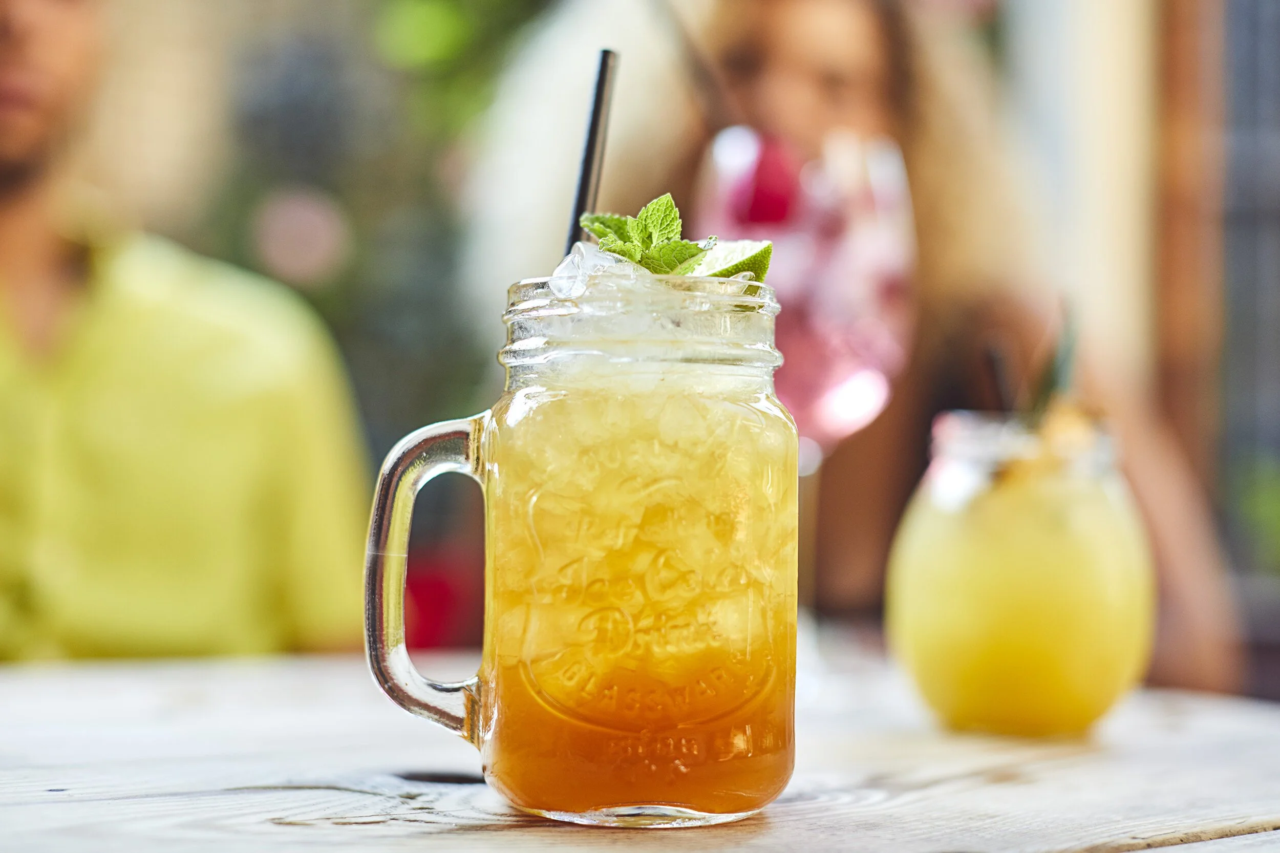 A mason jar glass with a yellow-orange beverage topped with ice, a lime wedge, and mint leaves, on a wooden surface, with blurred background of two people and another similar drink.