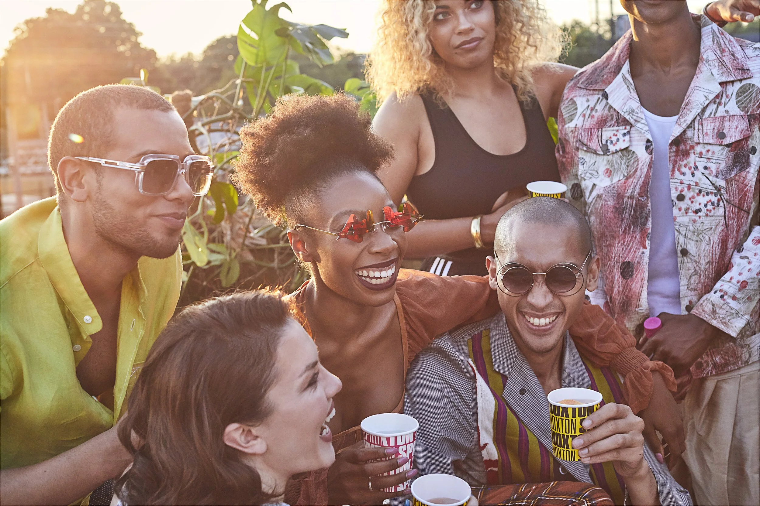 Group of diverse friends enjoying a sunny outdoor gathering, sitting and standing close together, smiling, holding cups, with greenery and sunset in the background.