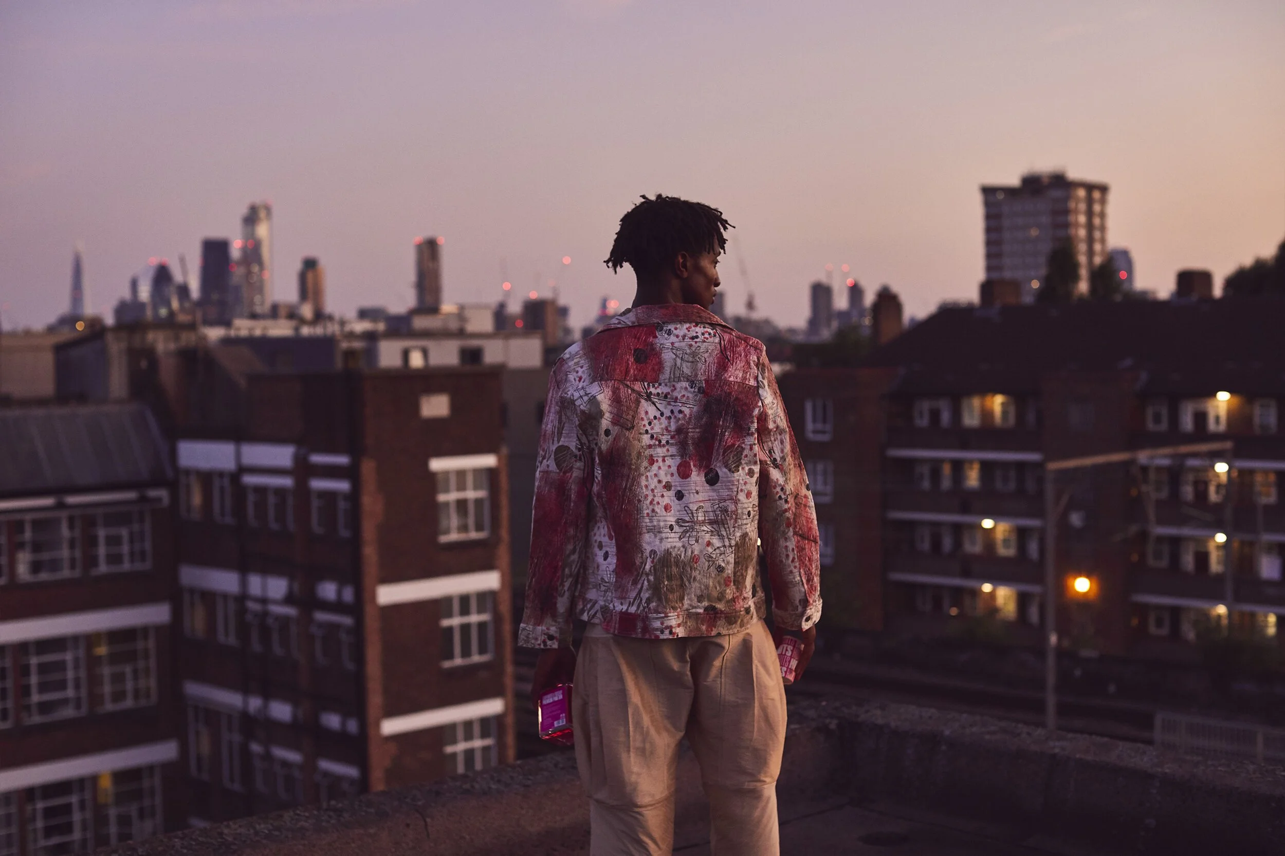 A person with short dreadlocks wearing a colorful abstract jacket and beige pants stands on a rooftop, looking over a city skyline at dusk, with high-rise buildings and residential blocks in the background.