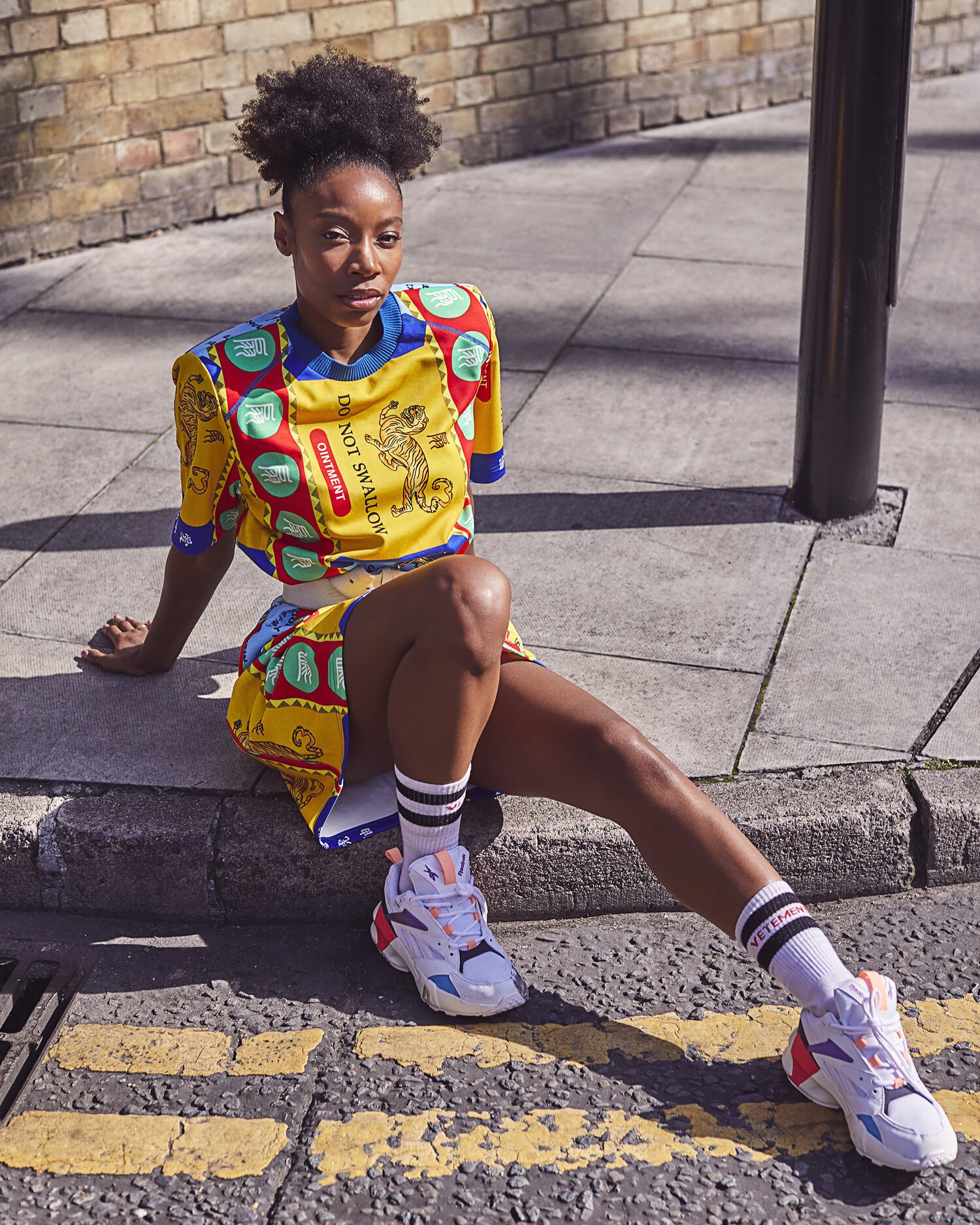 A woman sitting on a city sidewalk curb, dressed in colorful athletic wear, with sneakers, sitting near a pole and looking at the camera.