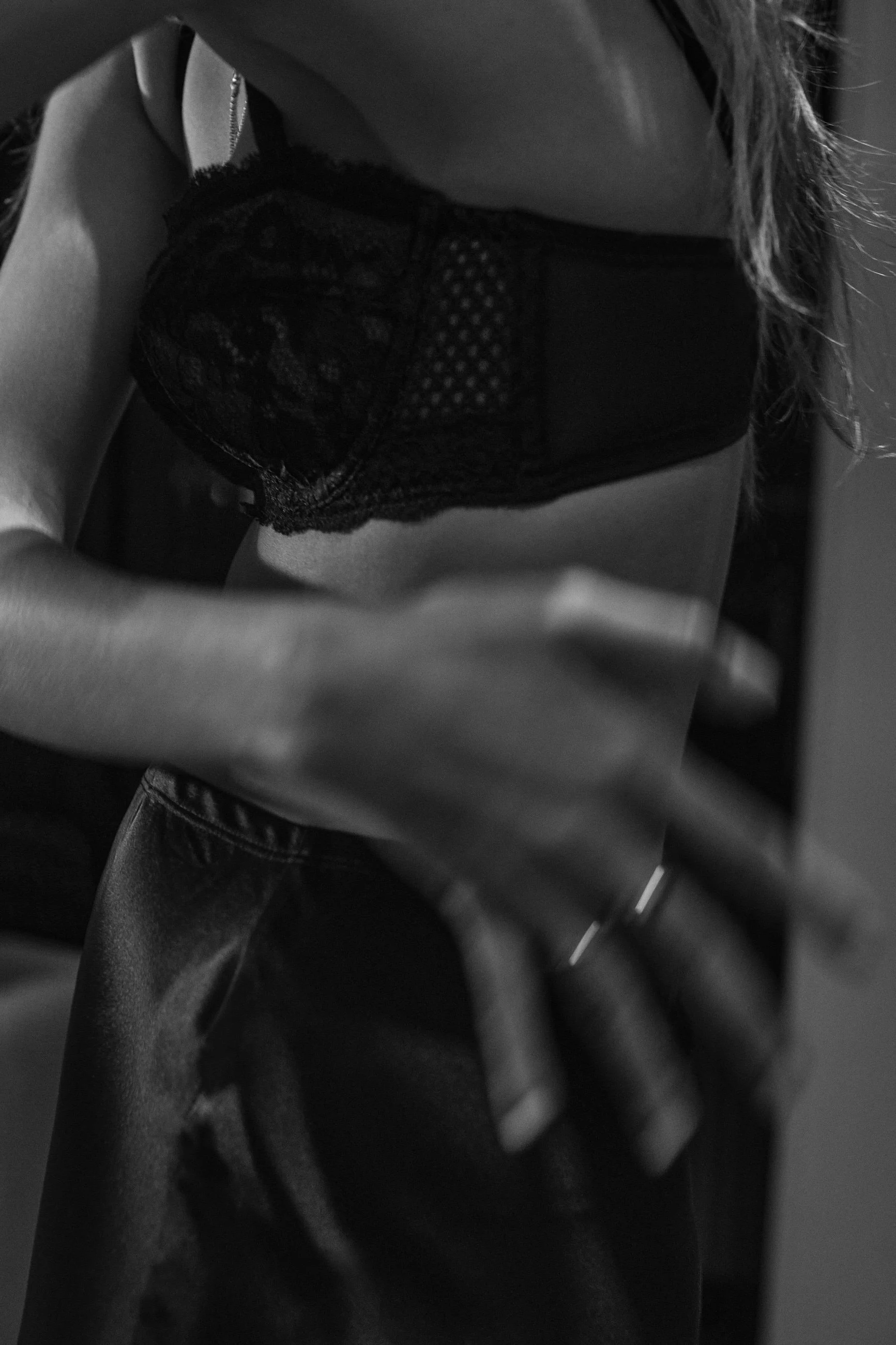 Close-up of a person wearing a lace and mesh black bra, taking a photo with a smartphone in black and white.