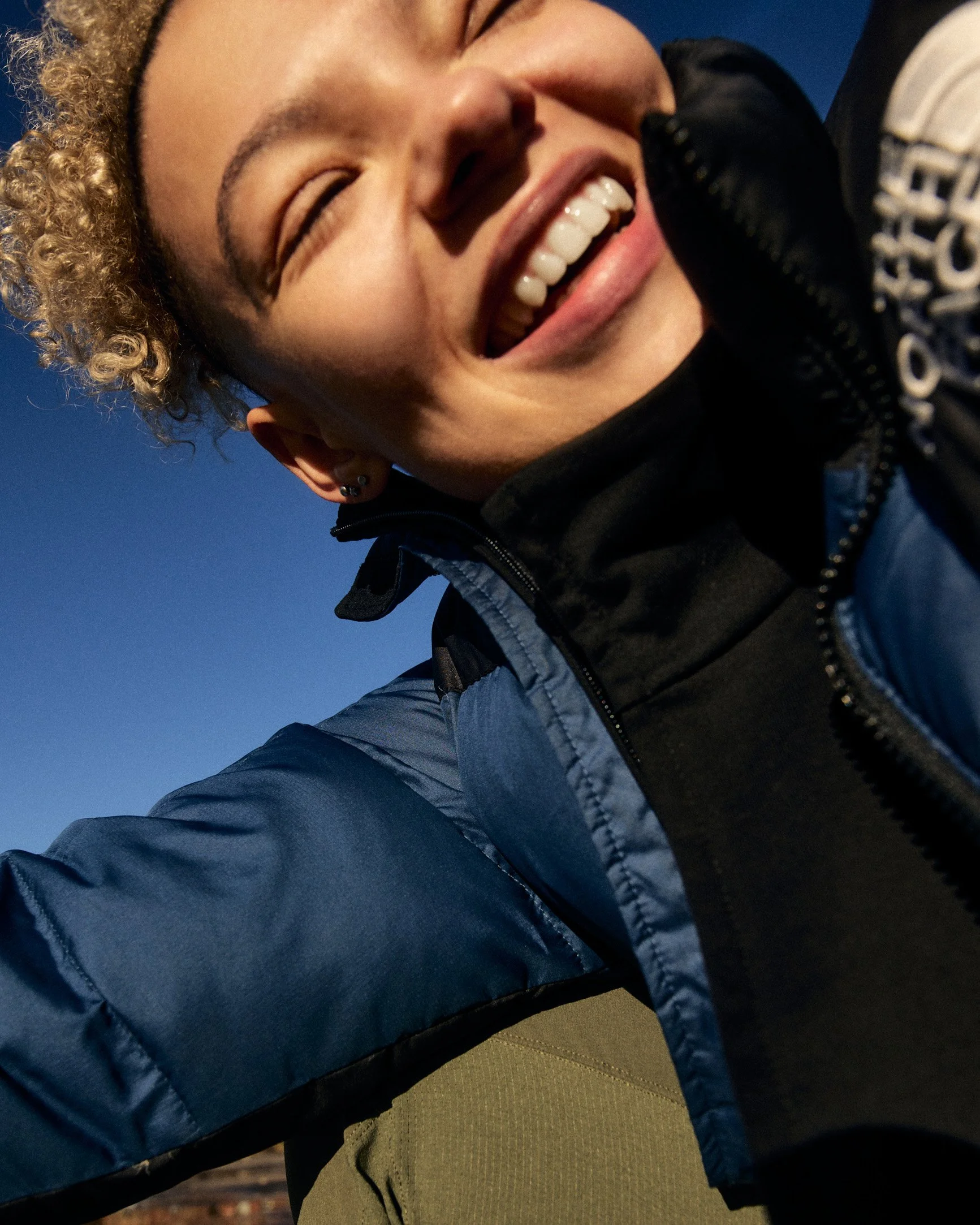 Close-up of a smiling person with curly blonde hair, wearing a black jacket with a logo, outdoors with a clear blue sky in the background.