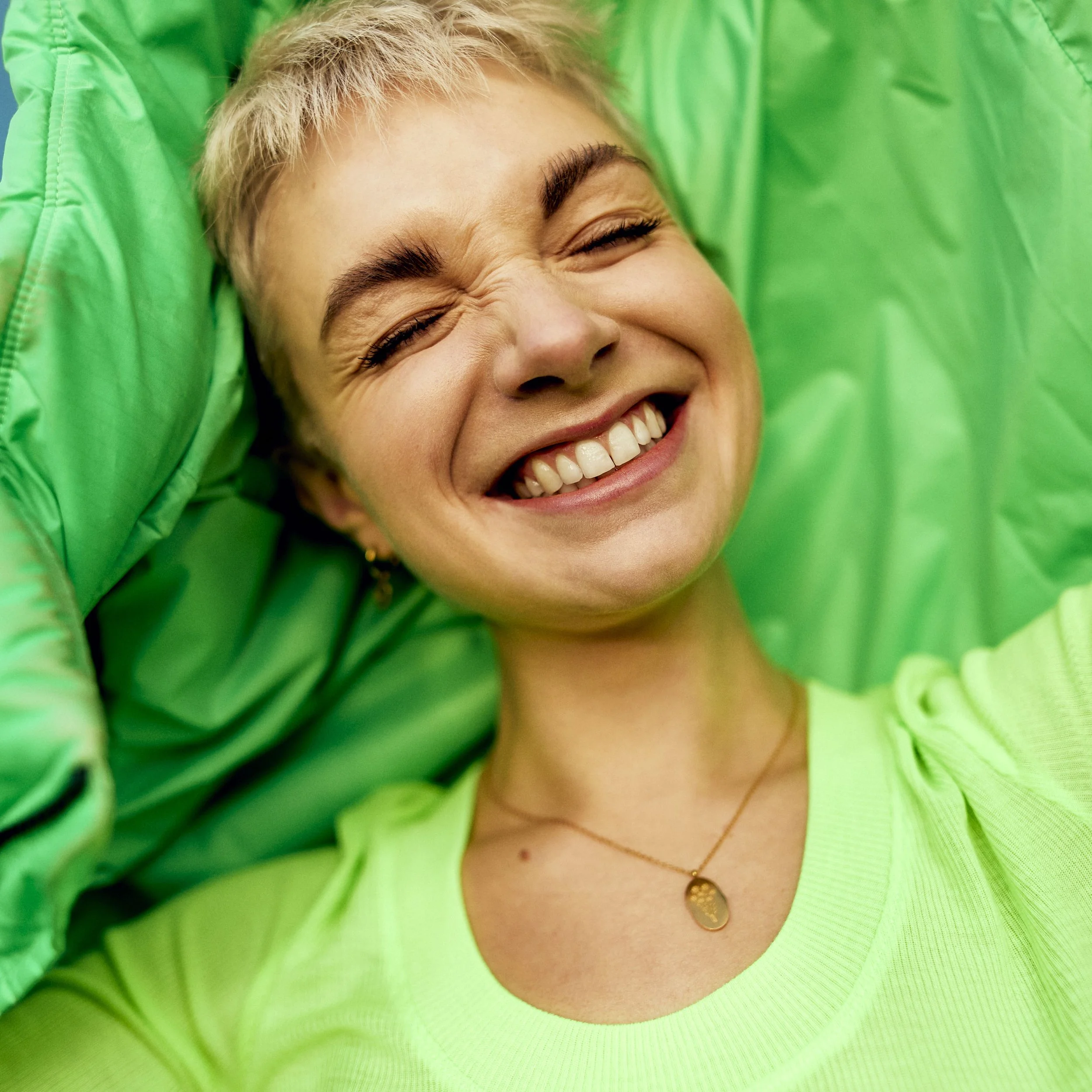 A young woman with short blonde hair smiling broadly with her eyes closed, surrounded by bright green clothing.