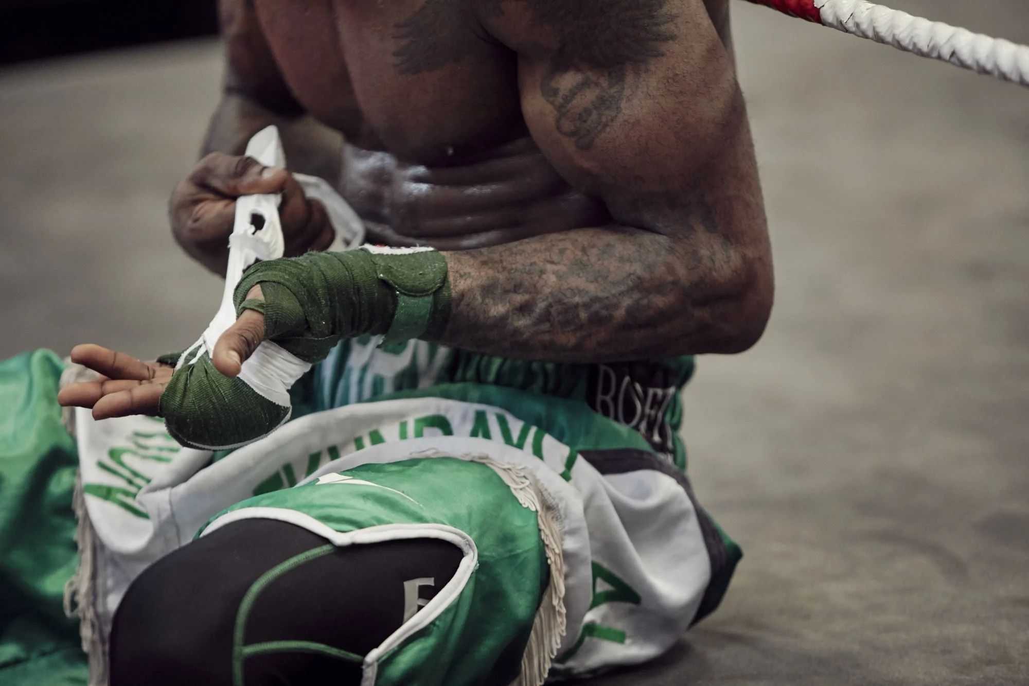 Close-up of a shirtless boxer with tattoos, sitting on the floor and wrapping his hands with green boxing tape.