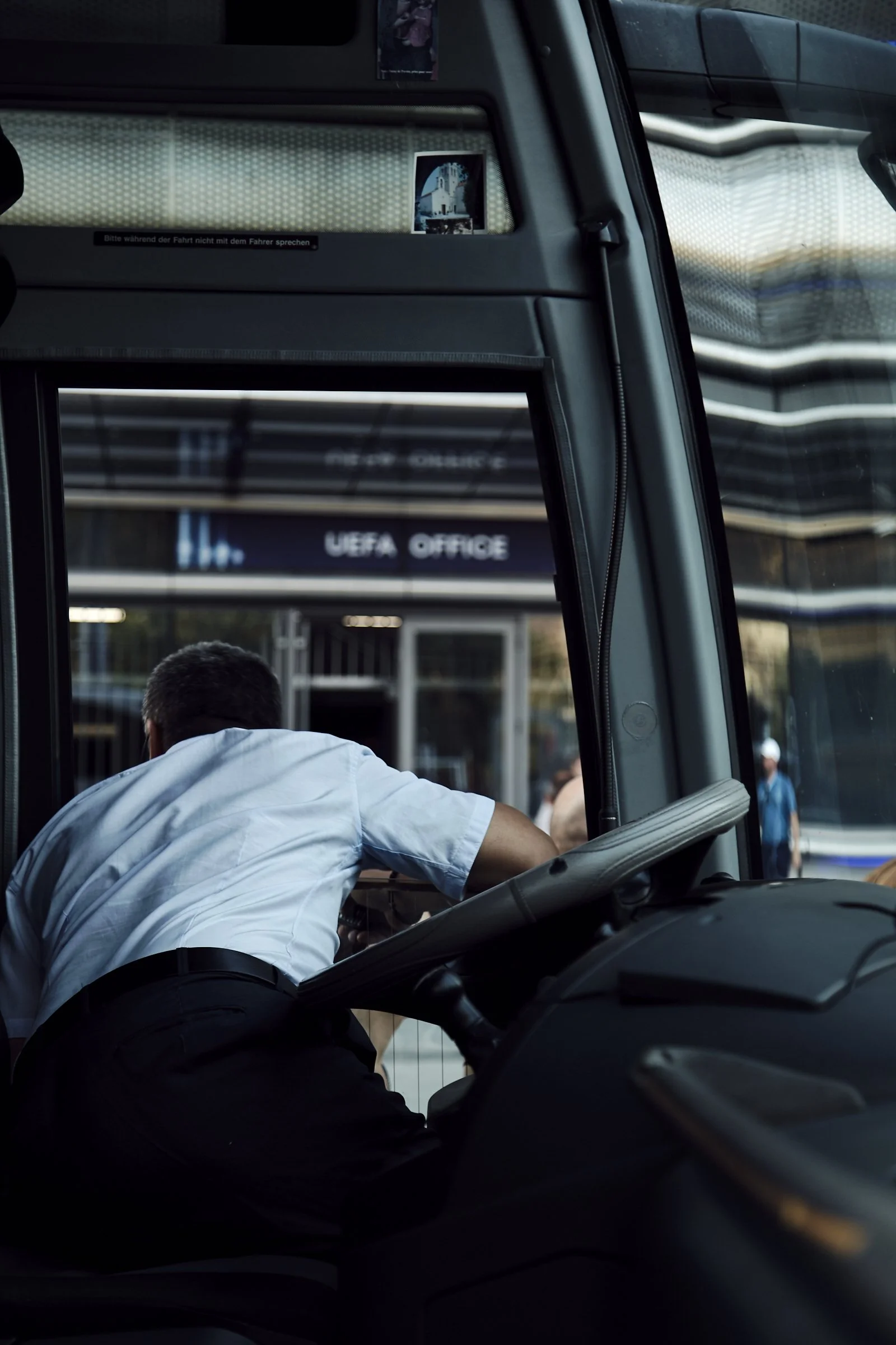 A man in a white shirt leaning over the dashboard of a bus, with an UEFA Office sign visible outside the window.