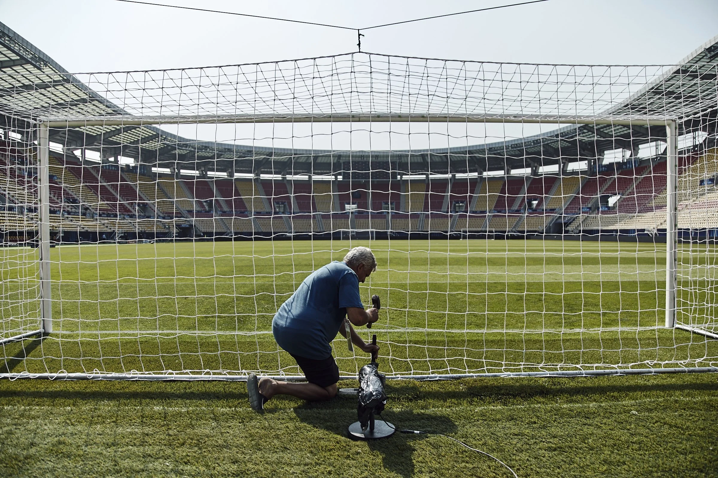 A man kneeling on the grass in front of a soccer goal, writing on a black bag. The stadium is empty with empty seats visible in the background.