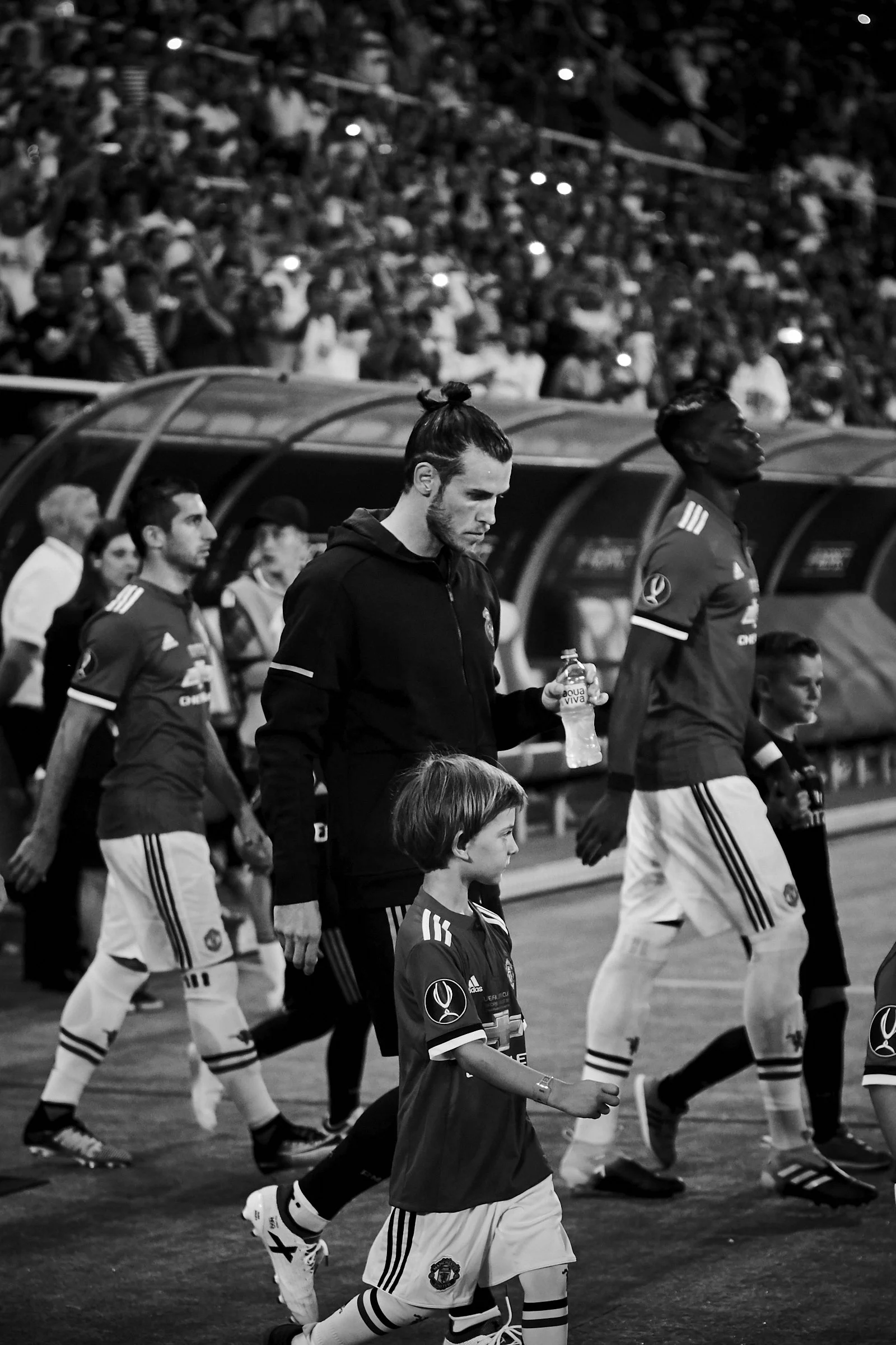 Black and white photo of soccer players and a young boy walking on the sidelines during a match, with a large crowd in the background.