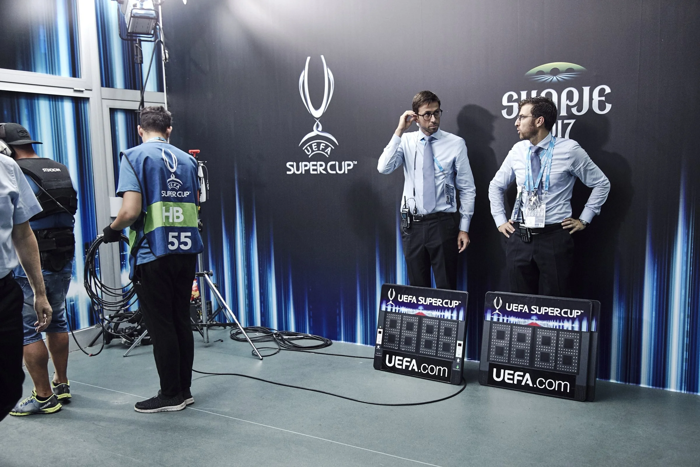 Event staff preparing for the UEFA Super Cup, with two men in dress shirts and ties standing against a wall with UEFA Super Cup logos, digital scoreboards on the floor, and team members handling cables and equipment.