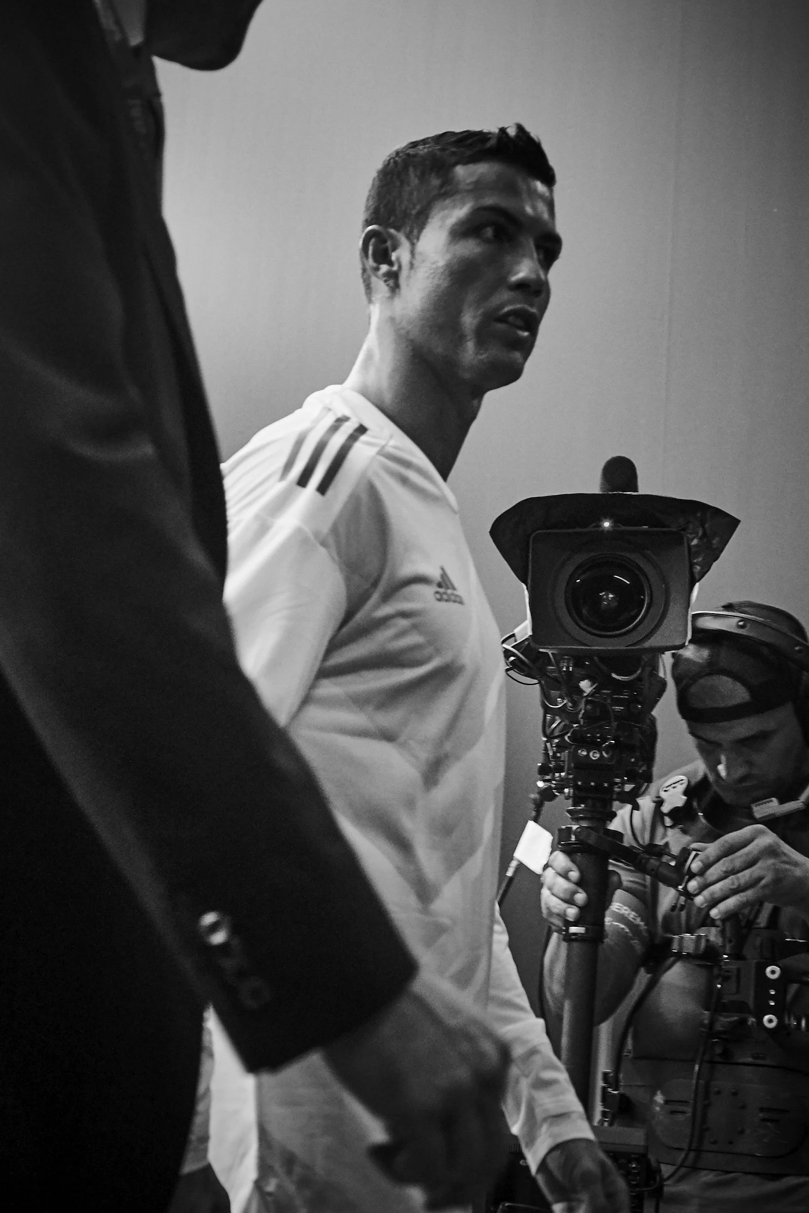 Black and white photo of Cristiano Ronaldo in a white Adidas jersey, being filmed or photographed in a professional setting, with technicians operating camera equipment around him.