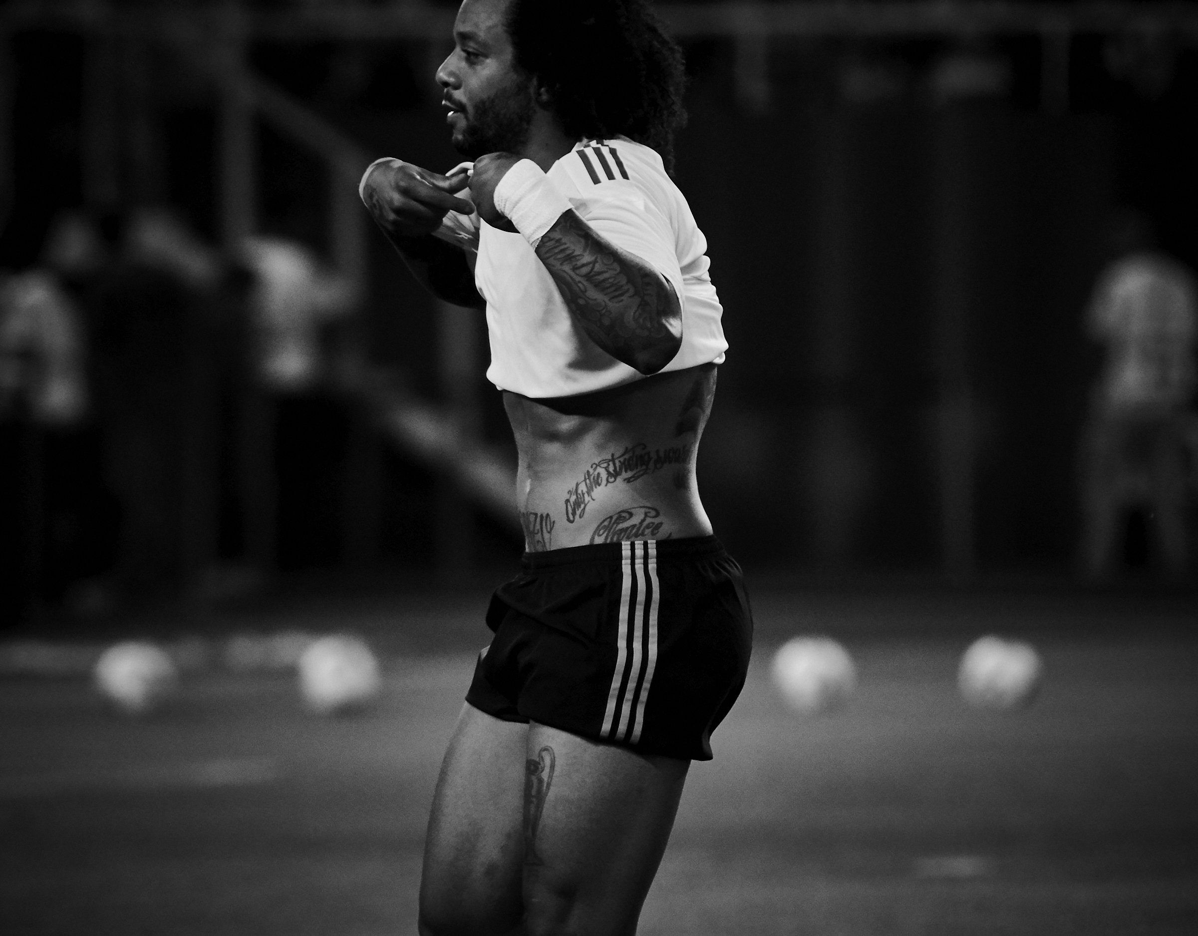 A man with tattoos, wearing a white shirt and black shorts, adjusting his shirt during a sports practice or game in a dimly lit indoor setting.