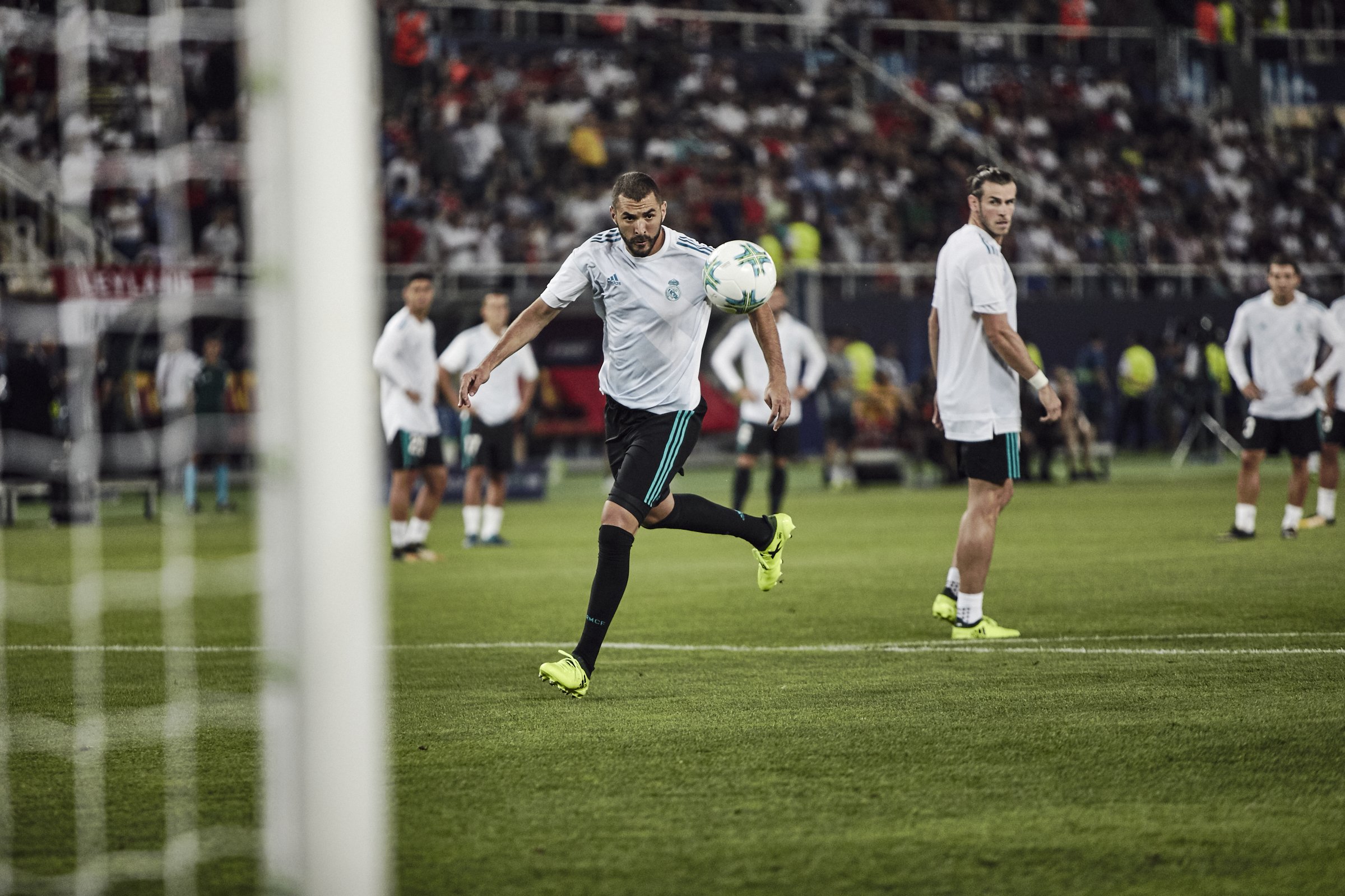 Soccer players warming up on a green field before a match, with a crowded stadium in the background.