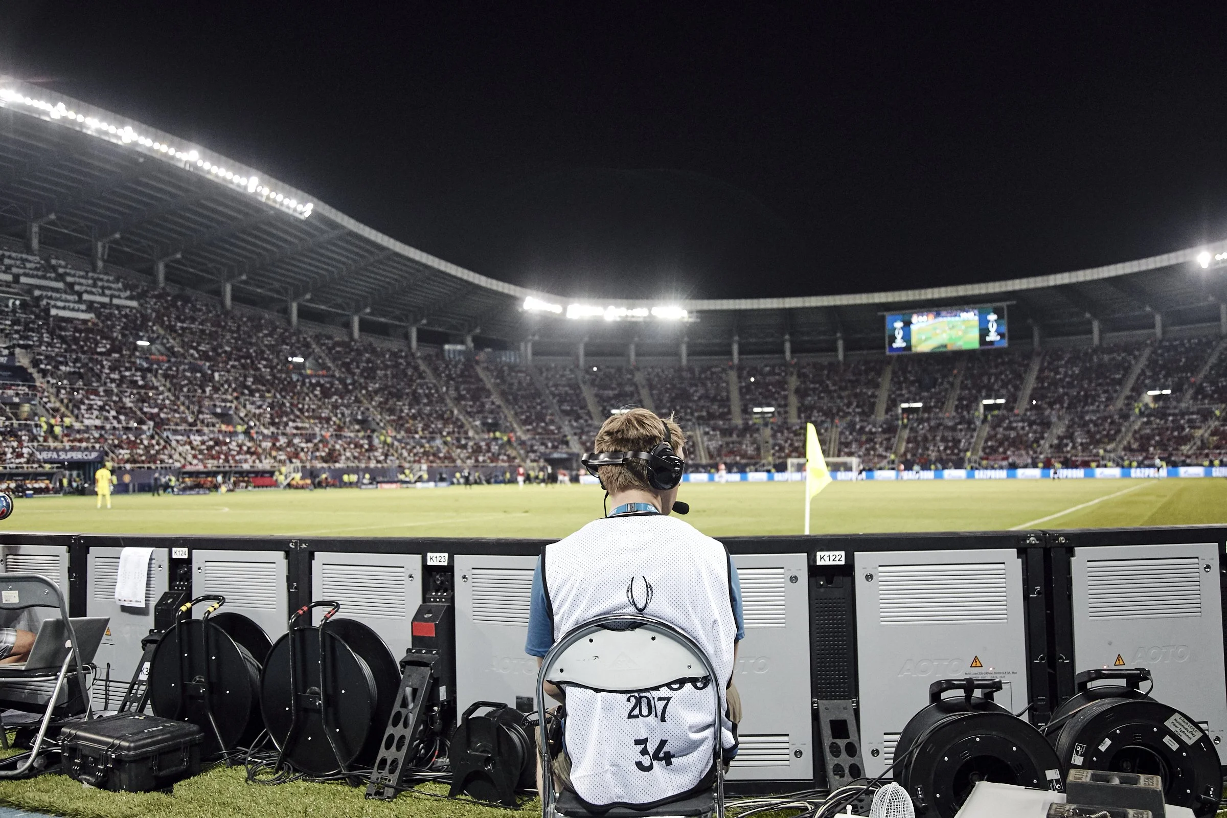 A sports commentator sitting in front of television broadcasting equipment at a soccer stadium during a night game. The stadium is filled with spectators, and a large screen displays the game.