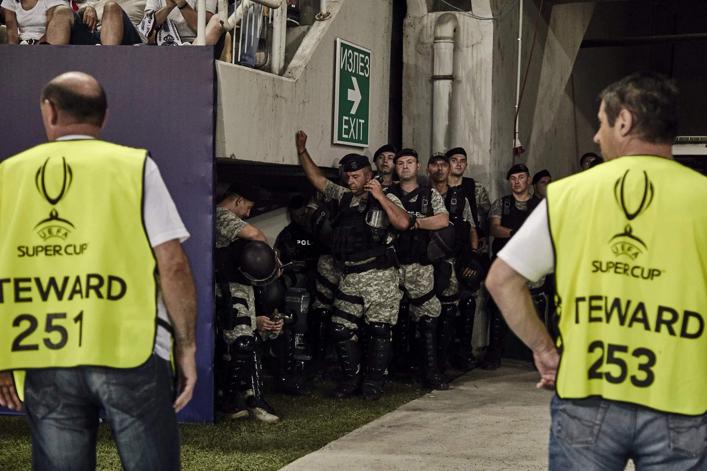 Security personnel in uniforms and protective gear standing and sitting against a concrete wall, with two men in yellow vests labeled 'STEWARD 251' and 'STEWARD 253' in the foreground, at an event or stadium.