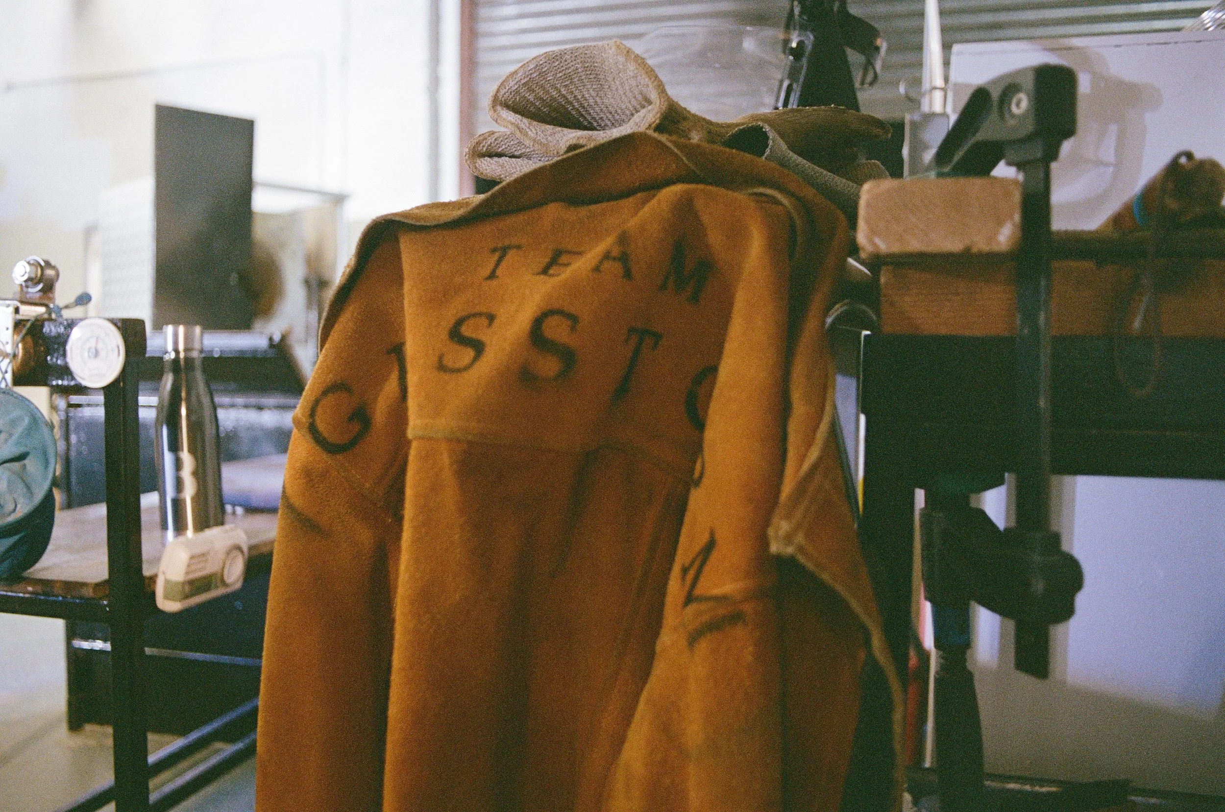 An orange leather jacket hanging on a black stand in a workshop, with the word 'GISS' visible on the back.