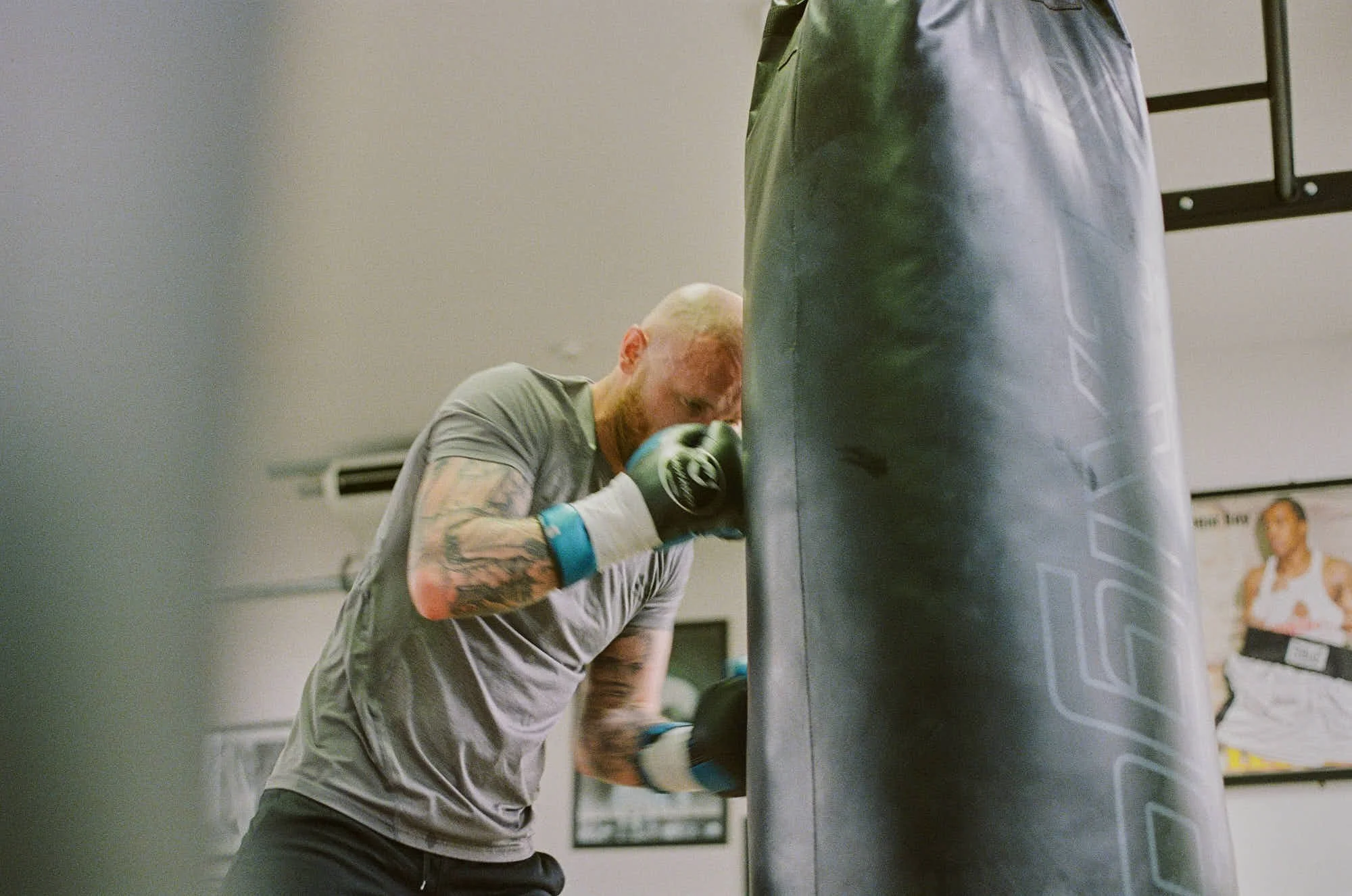 A man with tattoos and a beard wearing a gray shirt and boxing gloves punches a black heavy bag in a gym.
