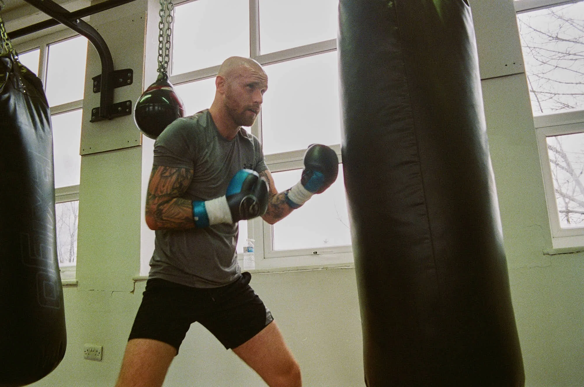 A man with tattoos is training with boxing gloves in a gym, practicing punches on a black punching bag.