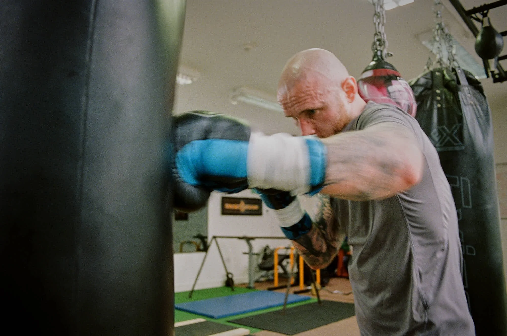 A boxer in gray shirt practicing punches on a black punching bag in a gym, wearing blue gloves and with tattoos on his left arm.