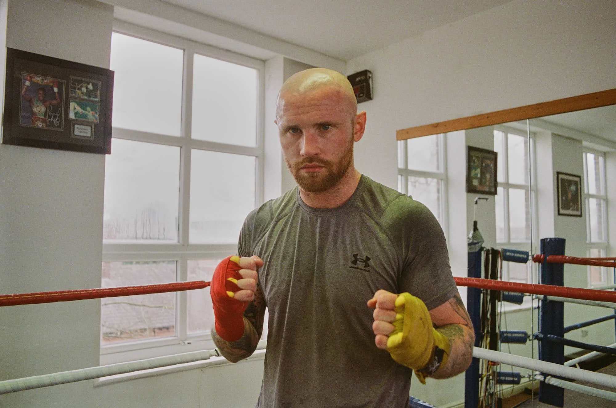 A male boxer with a shaved head and beard practicing in a boxing ring. He is wearing a gray Under Armour T-shirt and red and yellow hand wraps, standing in a fighting stance with fists raised. The boxing gym has large windows, framed pictures on the 