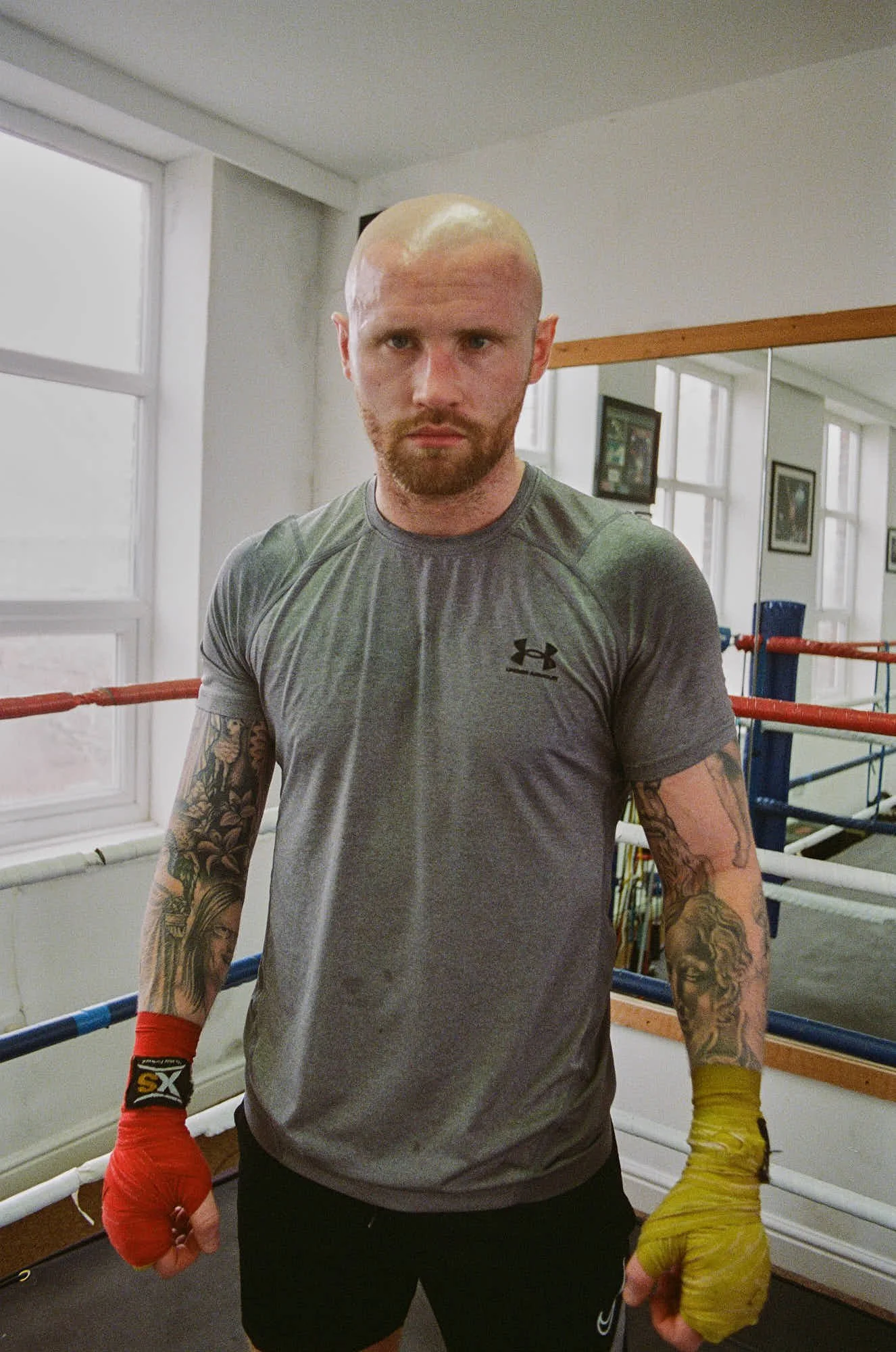 A man with a shaved head, beard, and tattoos on his arms, standing in a boxing gym inside a ring, wearing a gray Under Armour t-shirt, black shorts, and red and yellow hand wraps, looking directly at the camera.