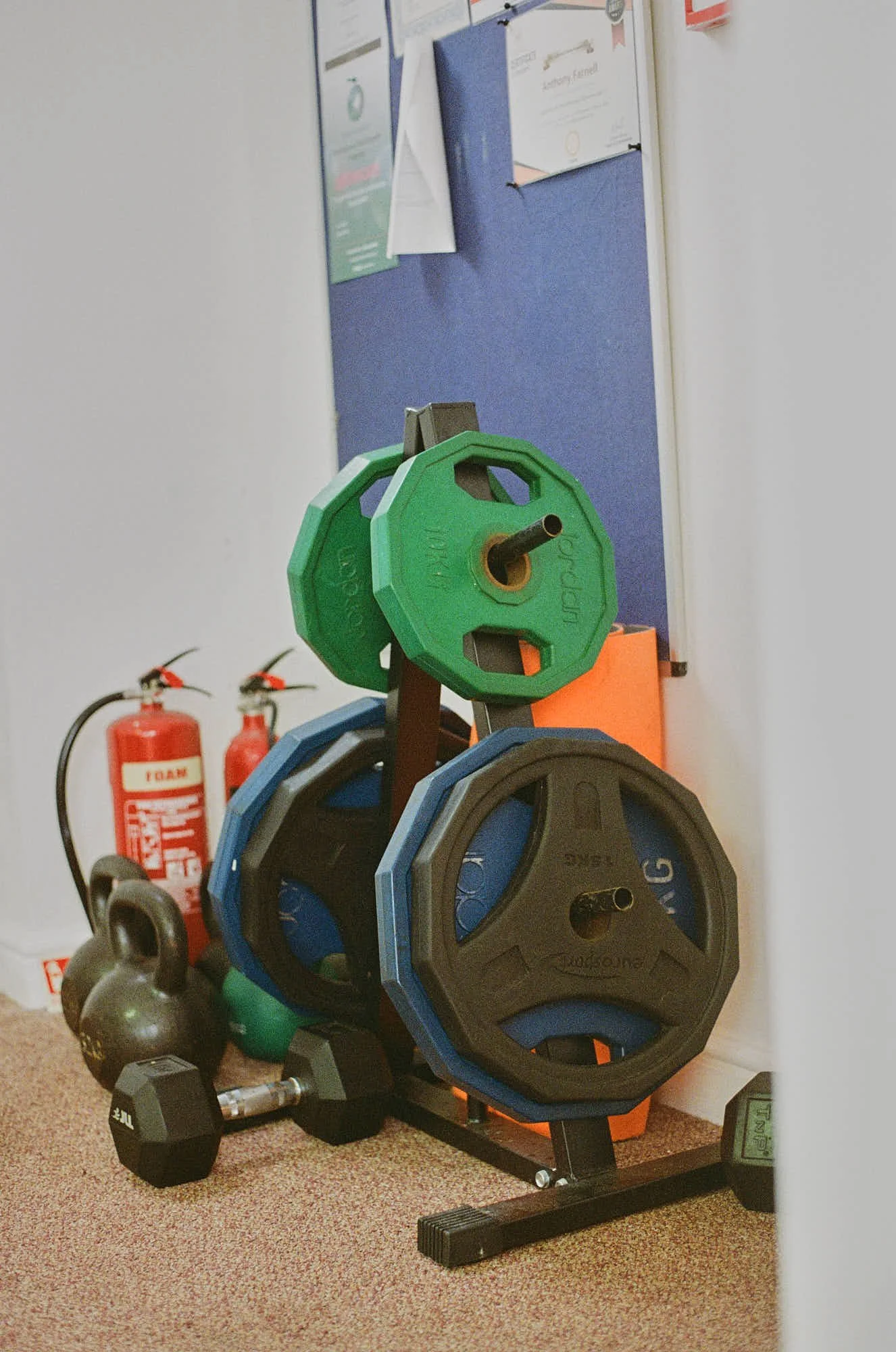 A workout area with colorful weight plates stacked on a rack, kettlebells on the floor, and fire extinguishers in the background.