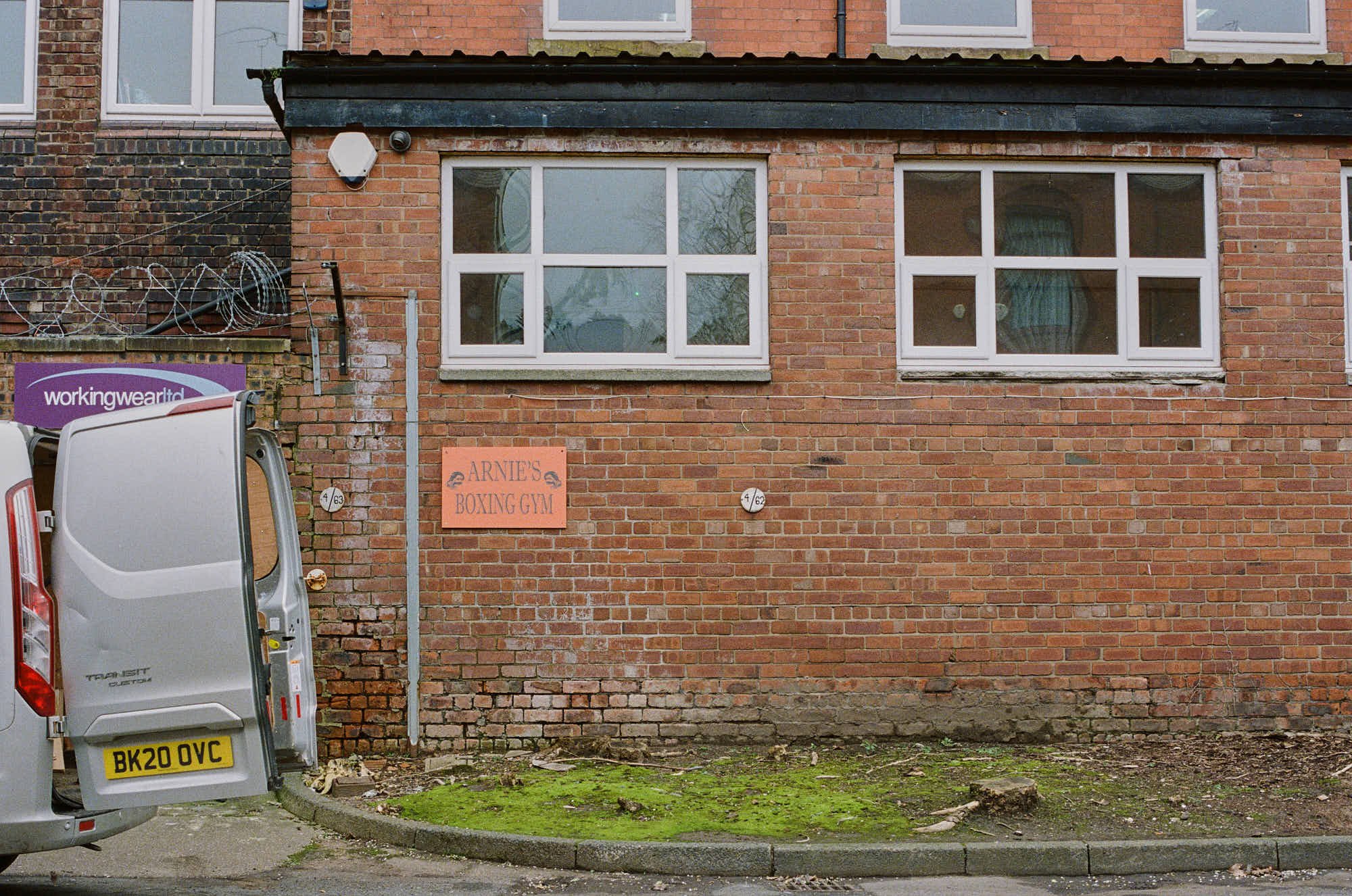 Brick building with four windows and a sign reading "Arnie's Boxing Gym" on the wall. A gray van with an open door is parked nearby, and there is green moss or grass on the ground.