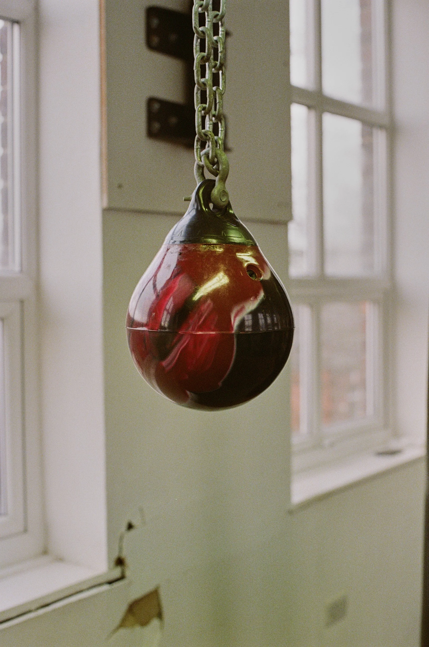 A glass pendulum with red and black sections hanging from a chain in a room with large windows.