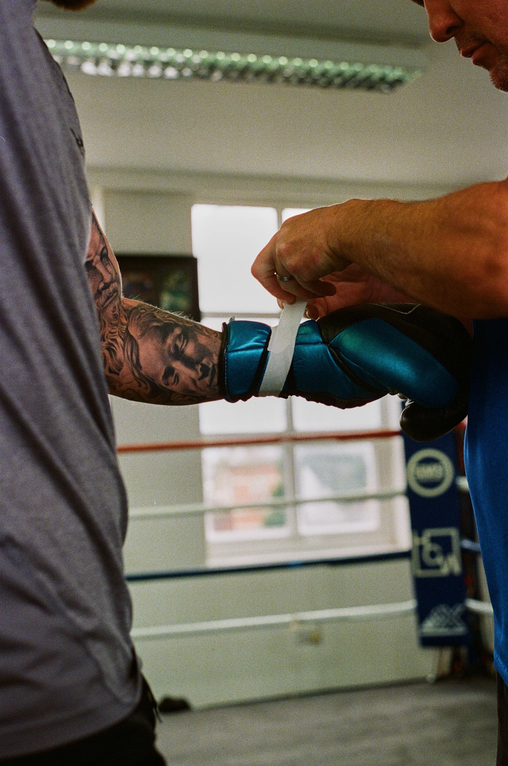 A boxer in gray clothing with tattoos on his arm is being fitted with boxing gloves by his coach or trainer in a boxing gym.