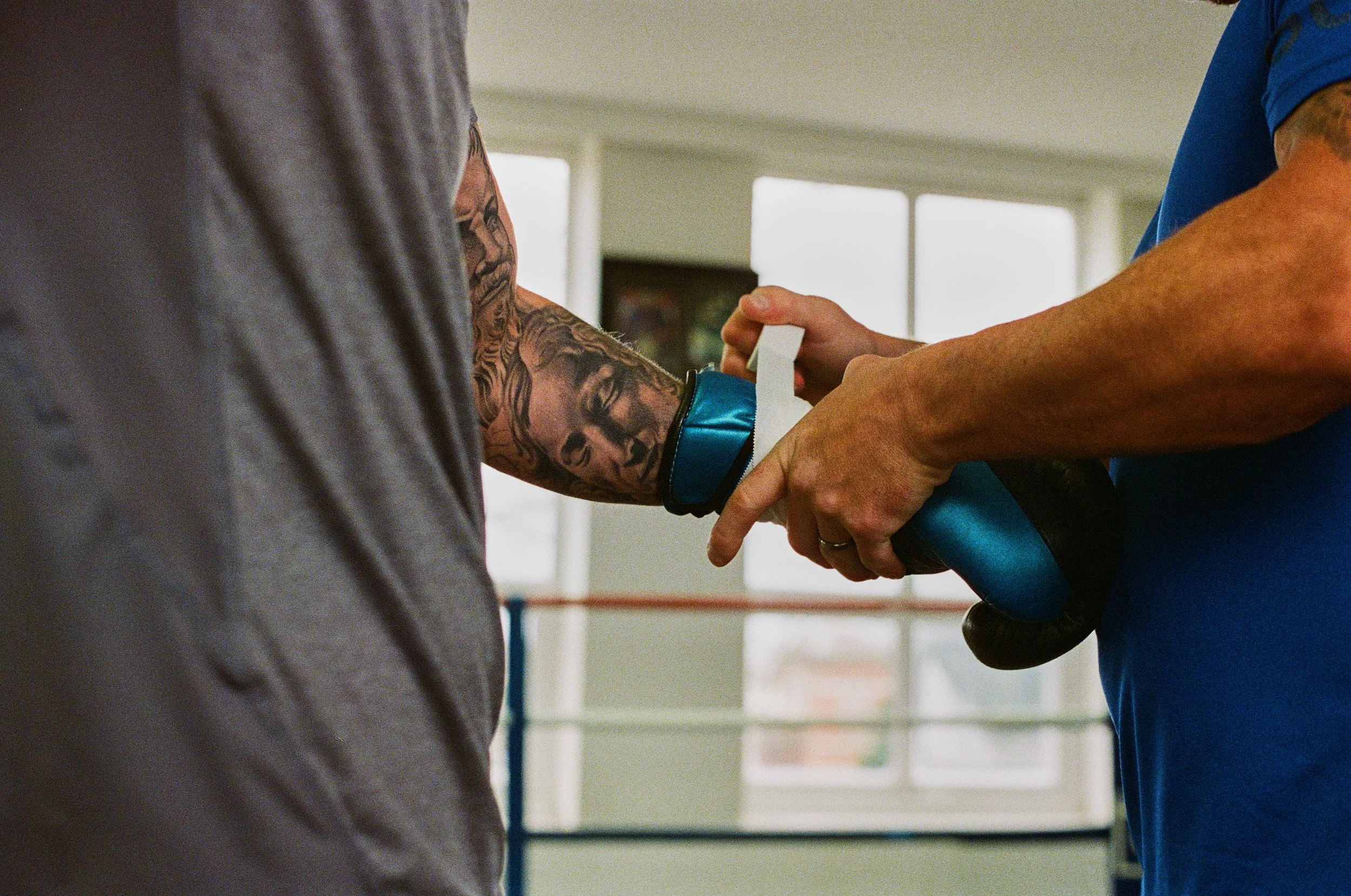 A person with a tattooed arm receiving a boxing hand wrap from another person, in a gym with windows and boxing rings in the background.