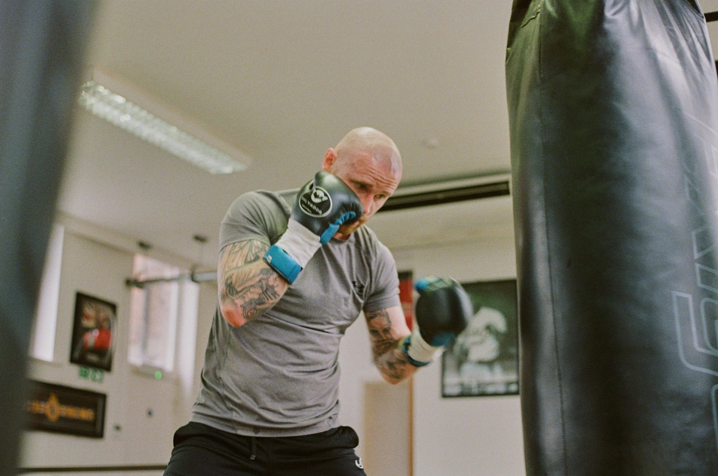 A male boxer with tattoos and a bald head practicing punches on a black punching bag in a gym.