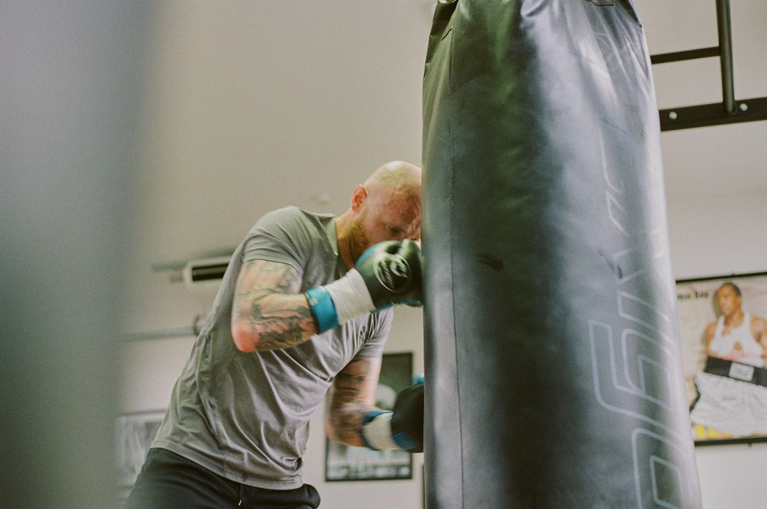 A man with tattoos and a bald head is practicing boxing in a gym. He is wearing boxing gloves and is hitting a punching bag.