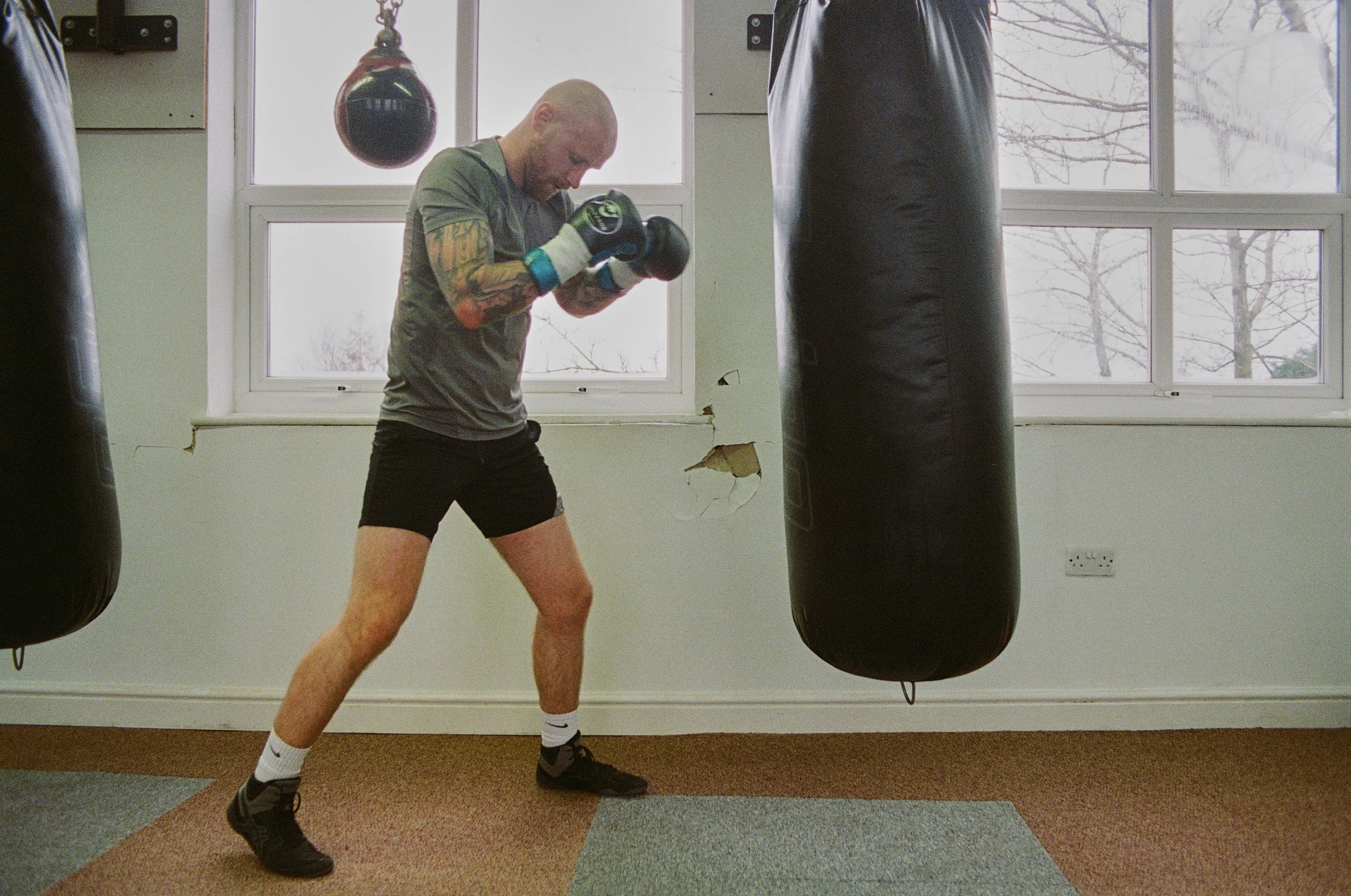 Man with tattoos wearing grey t-shirt, black shorts, black shoes, and boxing gloves in a boxing gym sparring with hanging black punching bags.