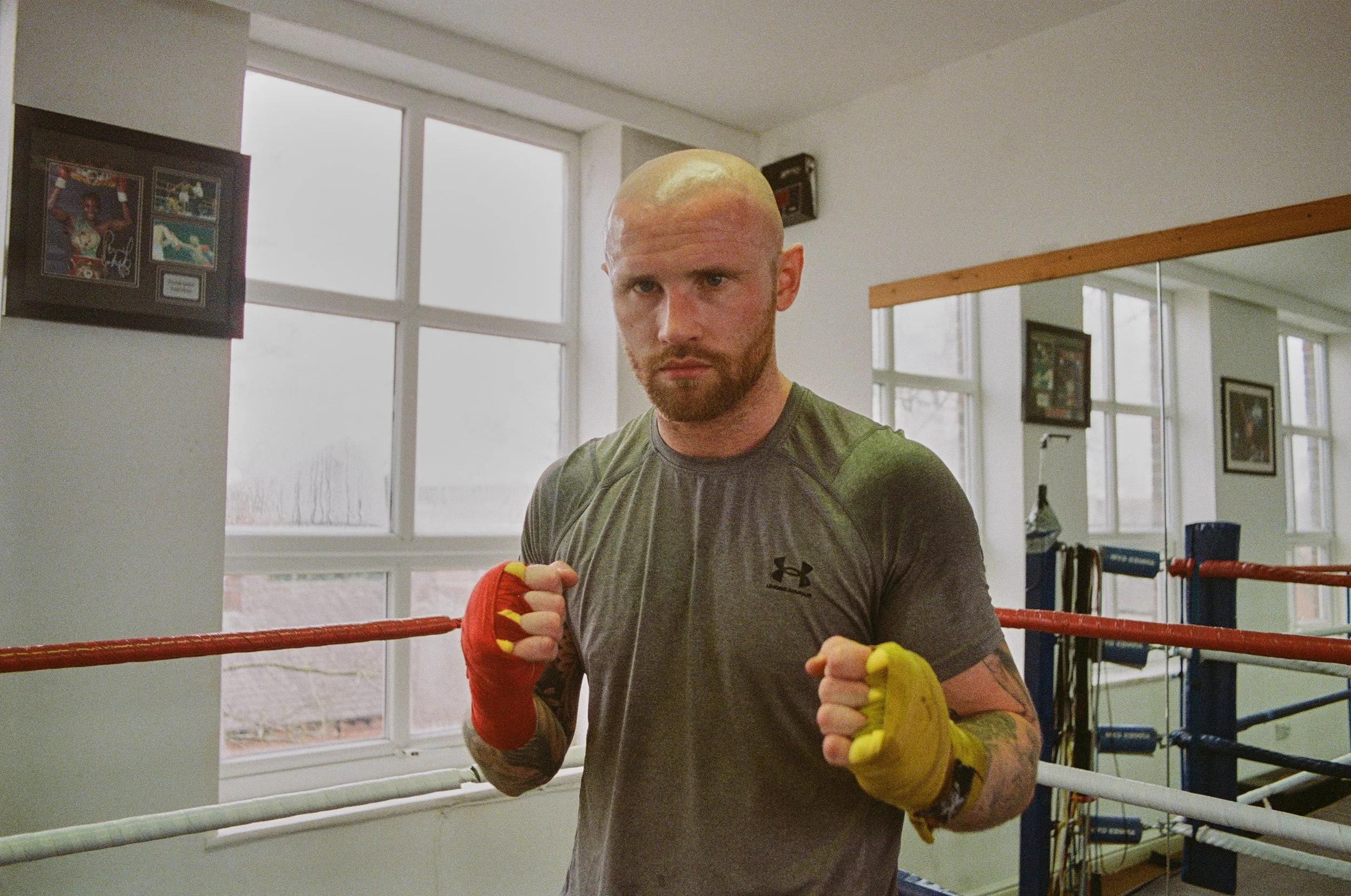 A male boxer inside a boxing ring, wearing boxing gloves and a gray Under Armour shirt, looking directly at the camera with a serious expression.