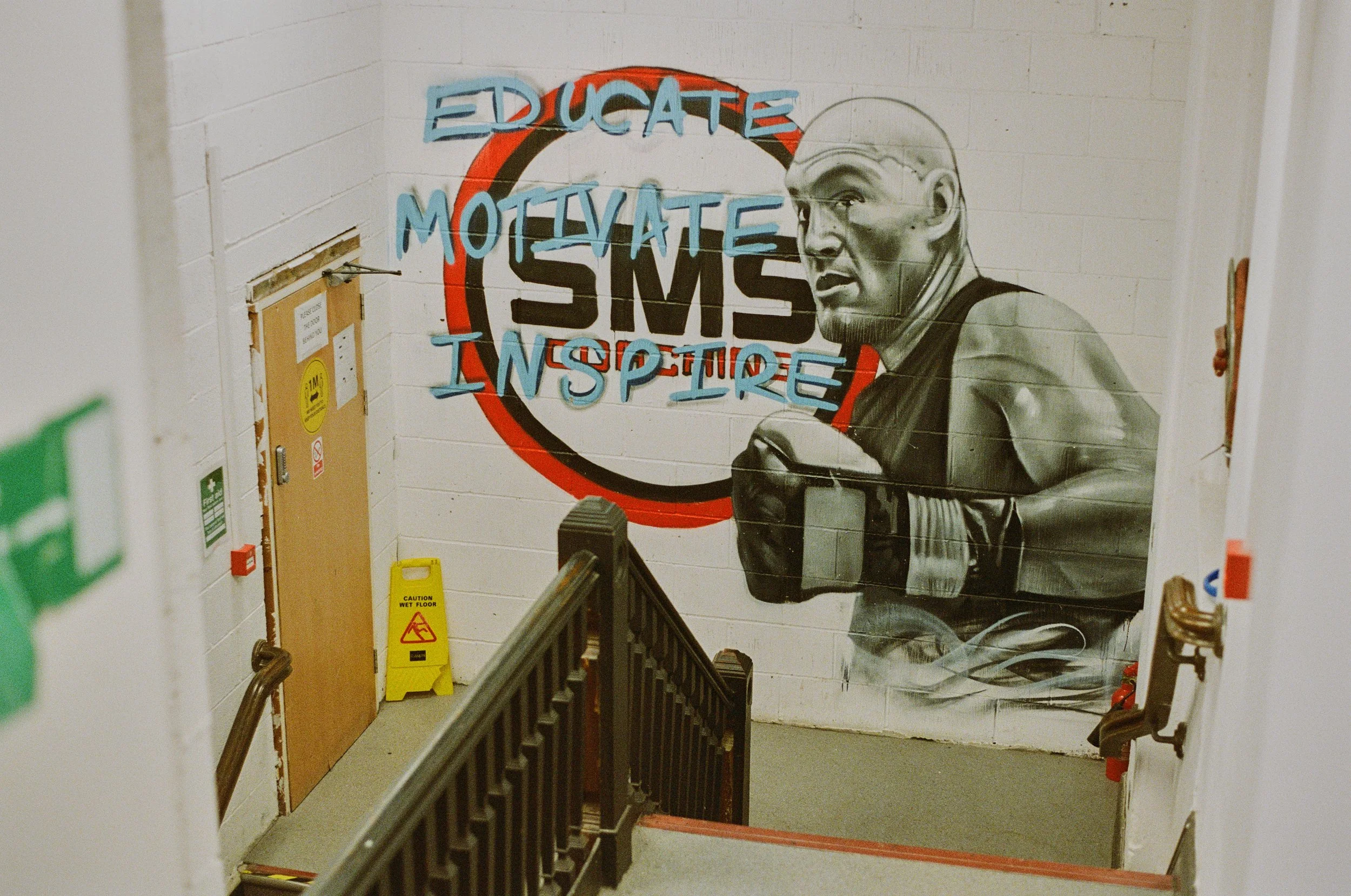 Graffiti mural of a muscular boxer with gloves, standing in front of a wall with the words 'EDUCATE MOTIVATE INSPIRE' in blue, above a stylized 'SMS' logo inside a red and black circle.