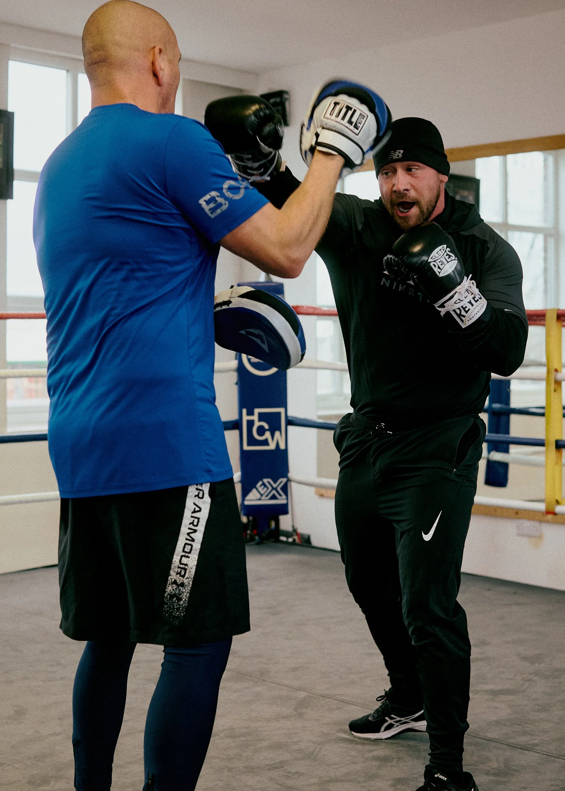 Two men boxing in a gym, one wearing a blue shirt and the other in black, practicing punches with boxing gloves.