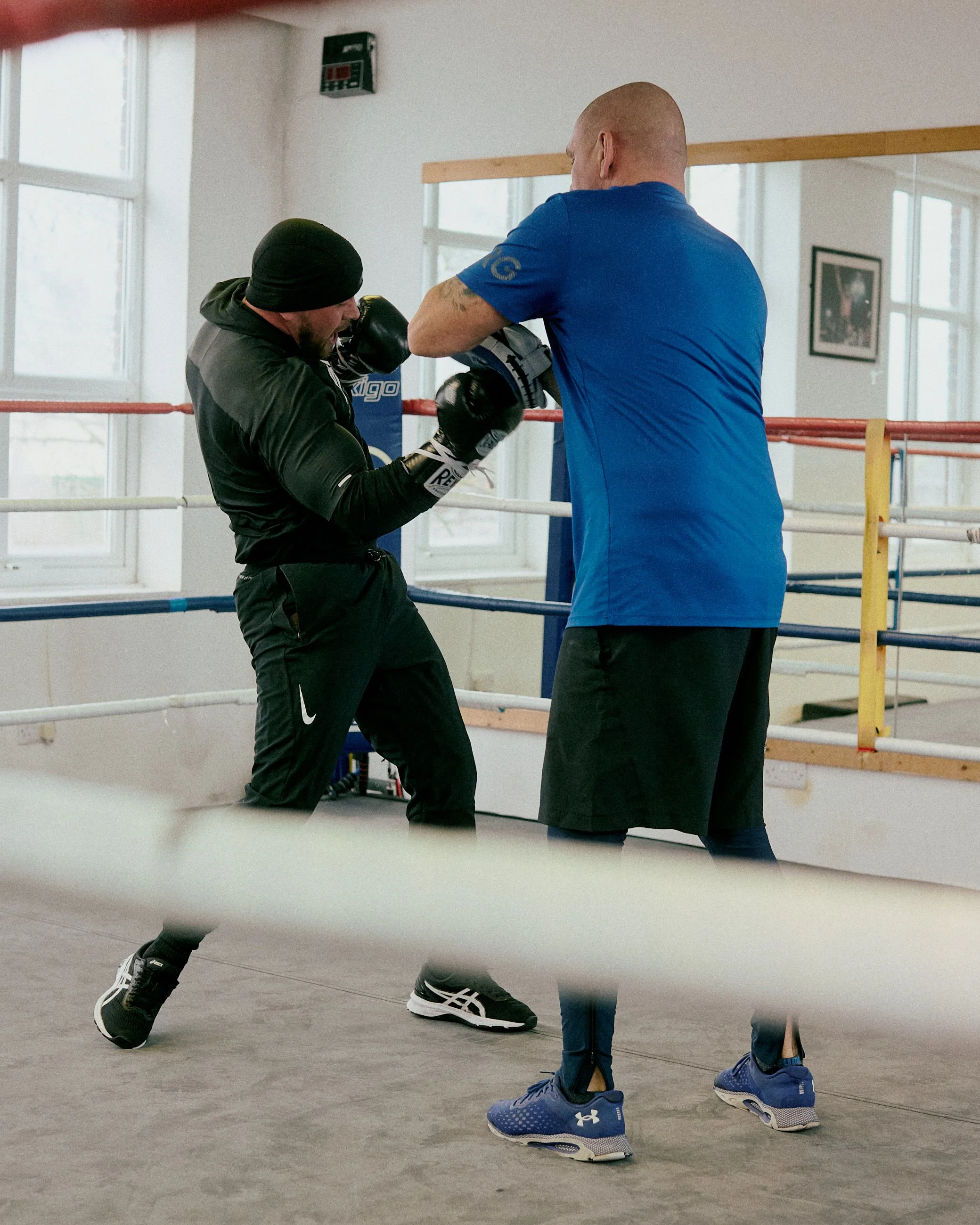 Two men practicing boxing in a gym, one with a black beanie and black jacket, the other in a blue shirt and shorts, inside a boxing ring with ropes and windows around.