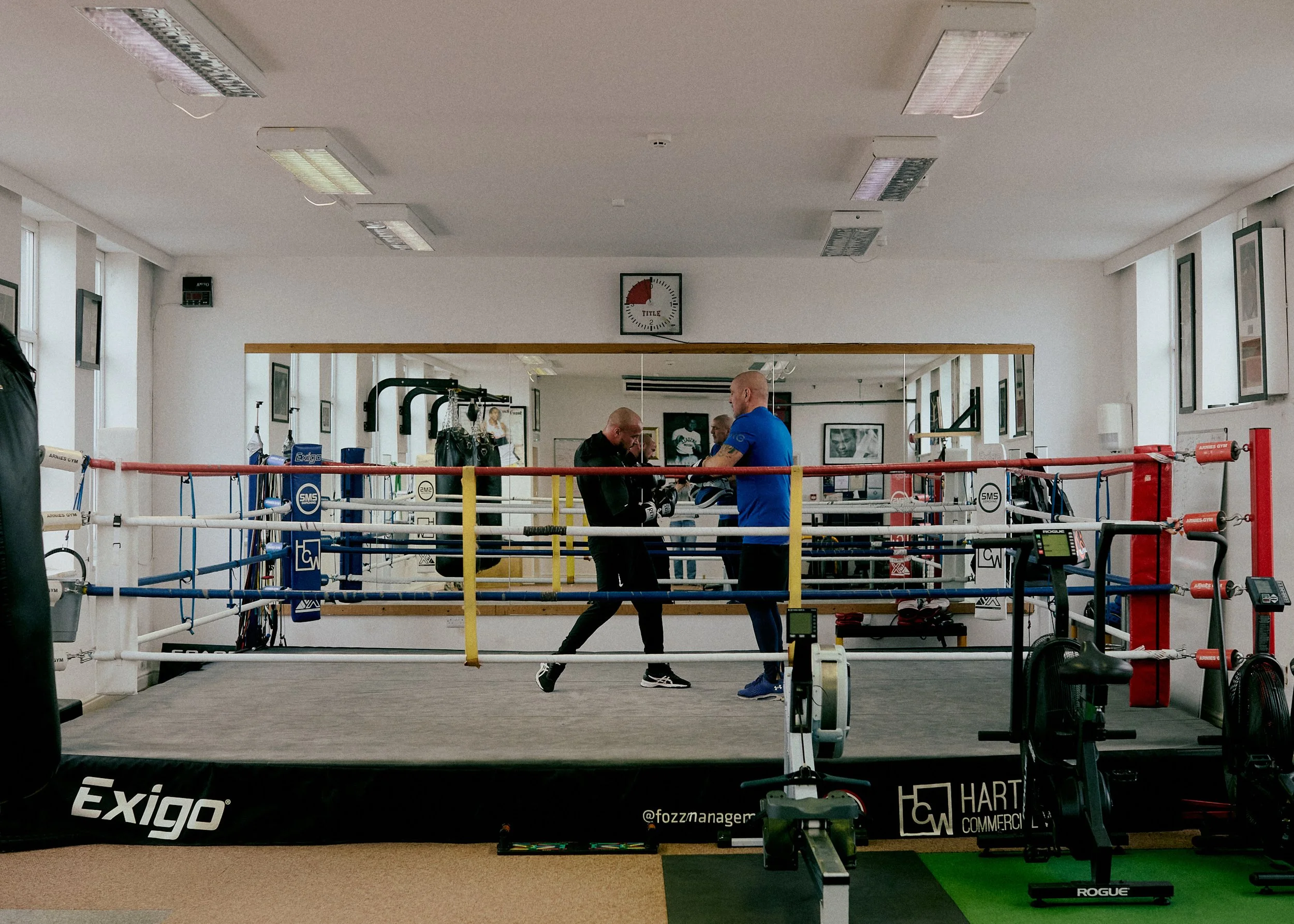 Two men training in a boxing gym with a boxing ring in the center, surrounded by gym equipment. One man appears to be showing or explaining something to the other.